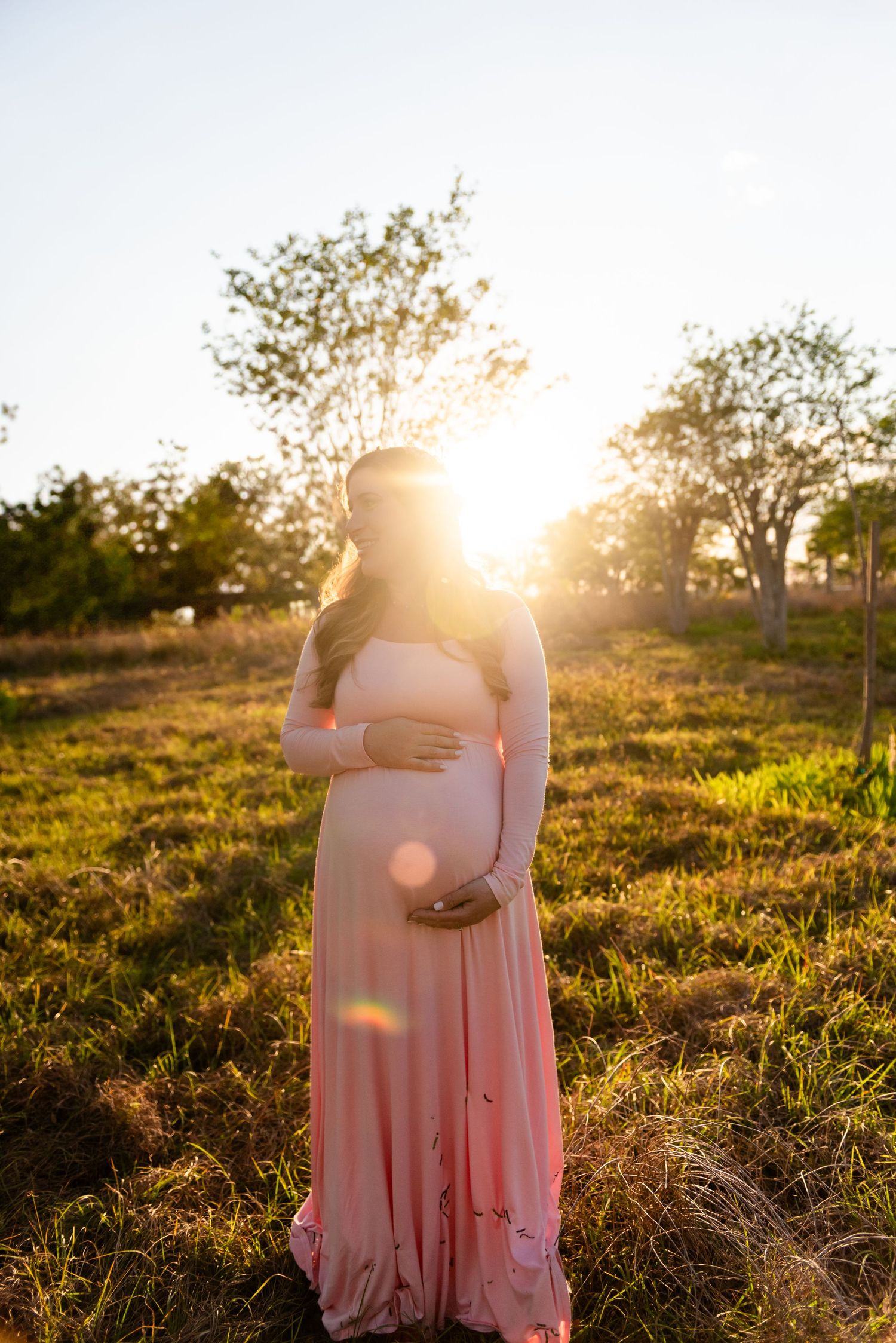 A dreamy maternity photoshoot at golden hour in a grassy field with soft sunflares and a pink flowy dress.
