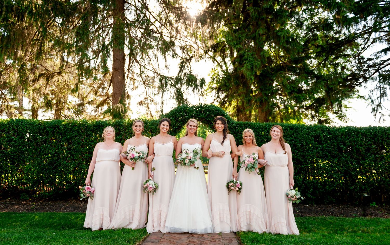 Wedding party in blush pink dresses poses against a backdrop of evergreen trees at sunset.
