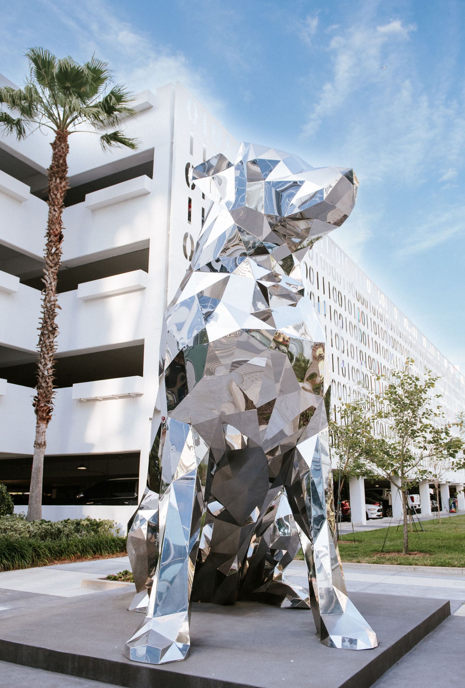 Large metallic geometric animal sculpture in front of a modern parking structure with palm trees.