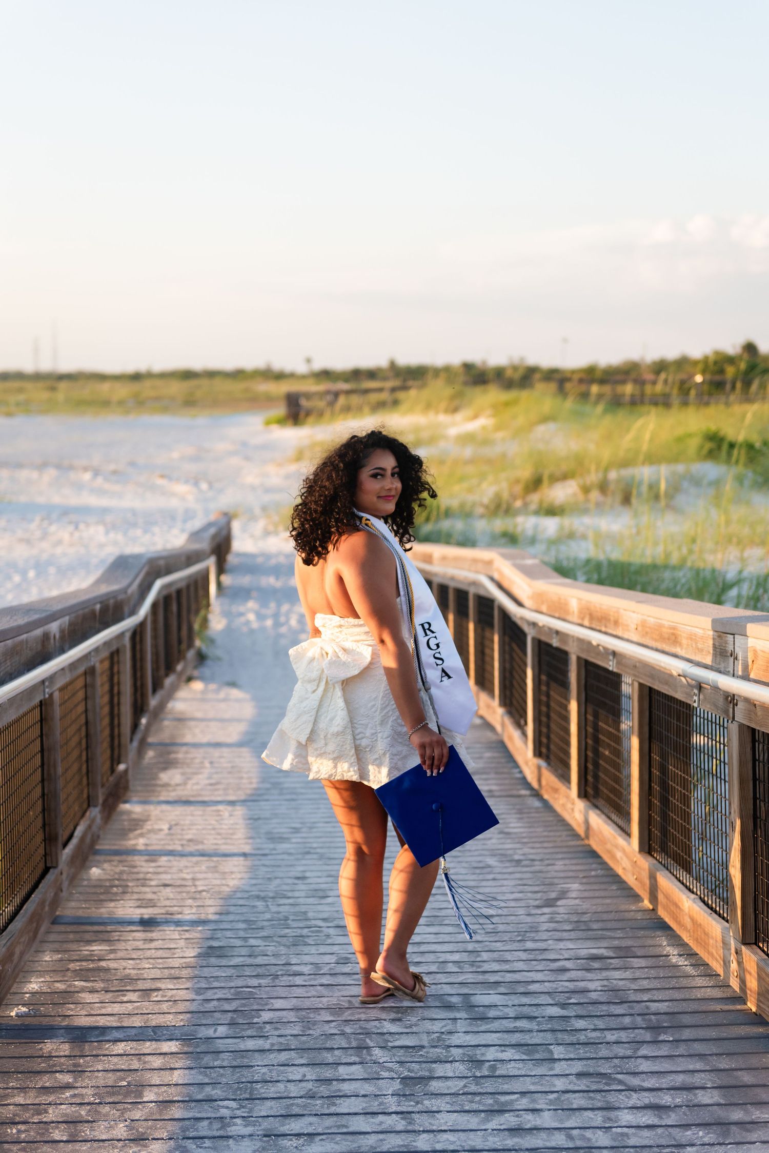 Person in white dress posing on wooden boardwalk with coastal scenery.