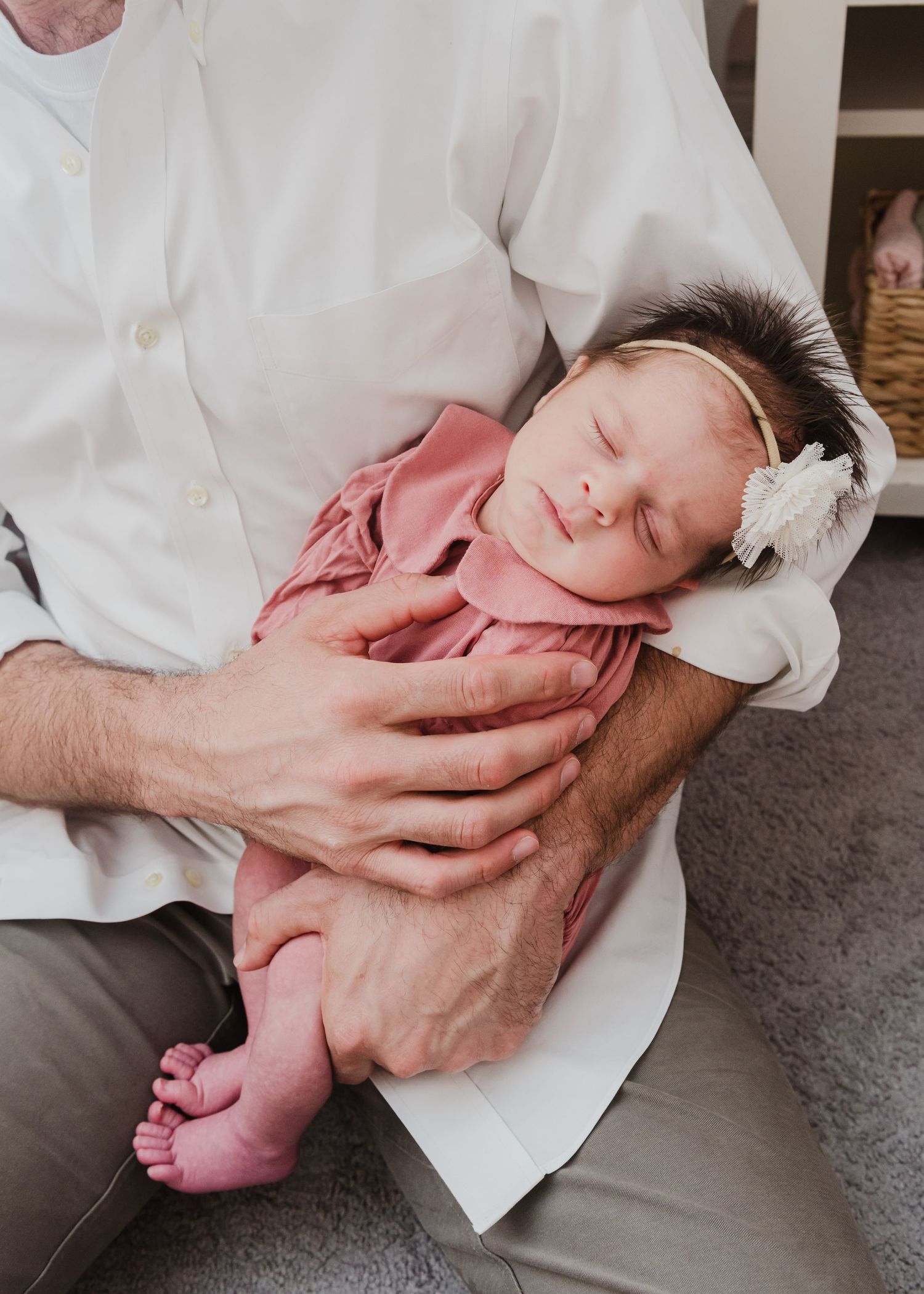 Sleeping newborn wearing pink outfit and white bow headband rests peacefully in parent's arms.