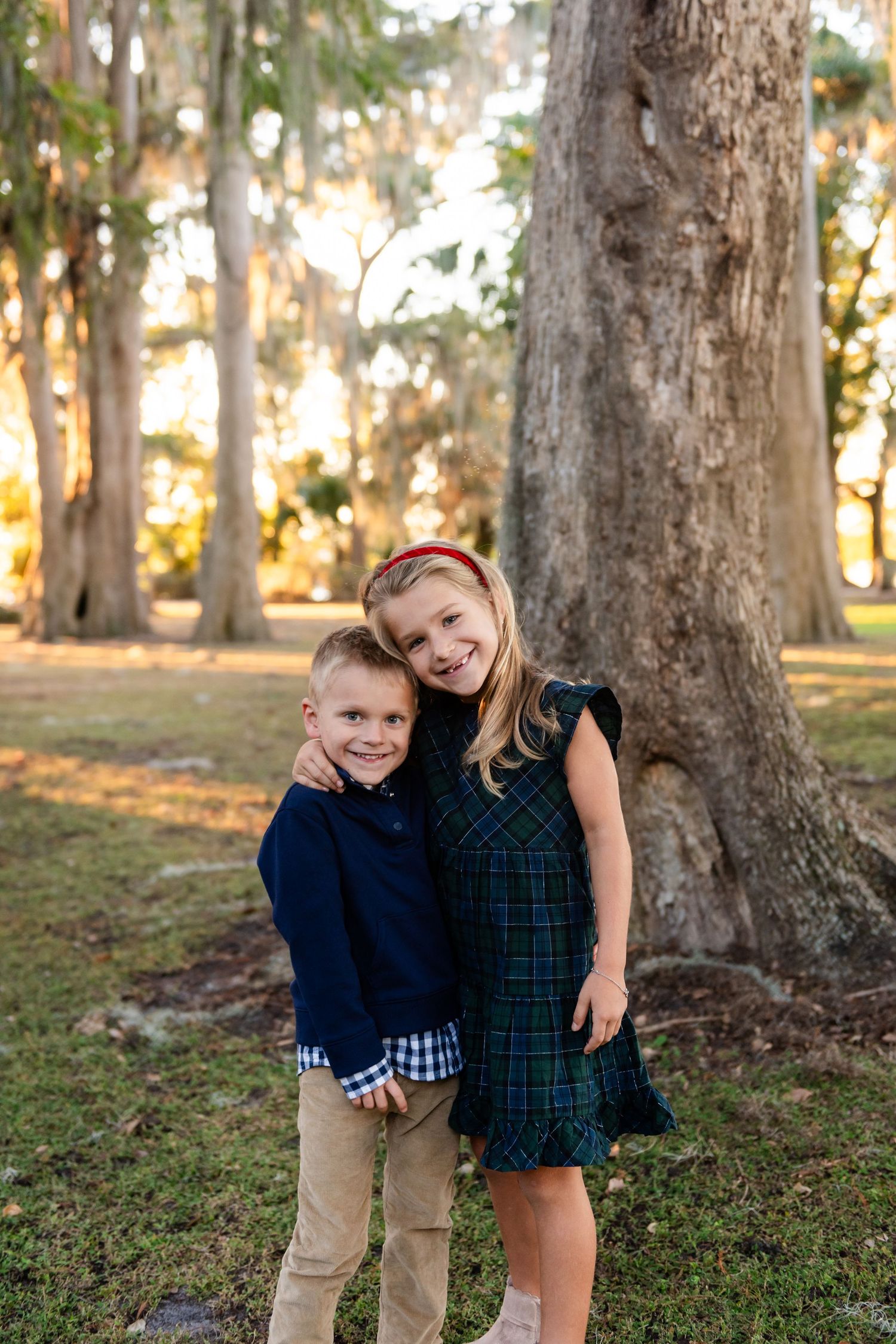 Two young children in classic preppy outfits stand together smiling beside a large tree trunk in an autumn park setting.