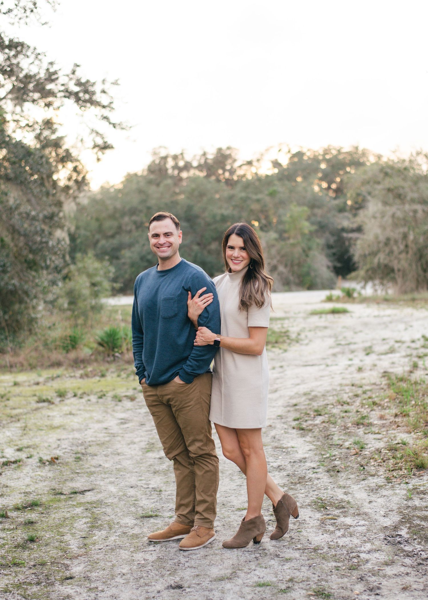 A couple stands together on a dirt path surrounded by trees at dusk, wearing casual attire and boots.