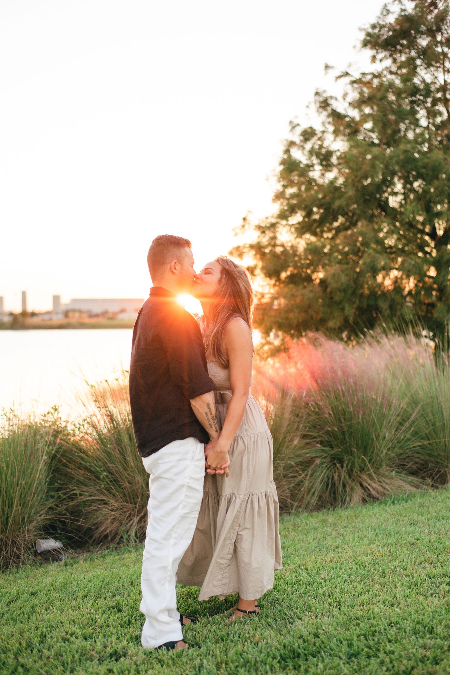 Couple embraces in romantic sunset moment by waterfront with ornamental grasses and golden sunlight glowing behind them.