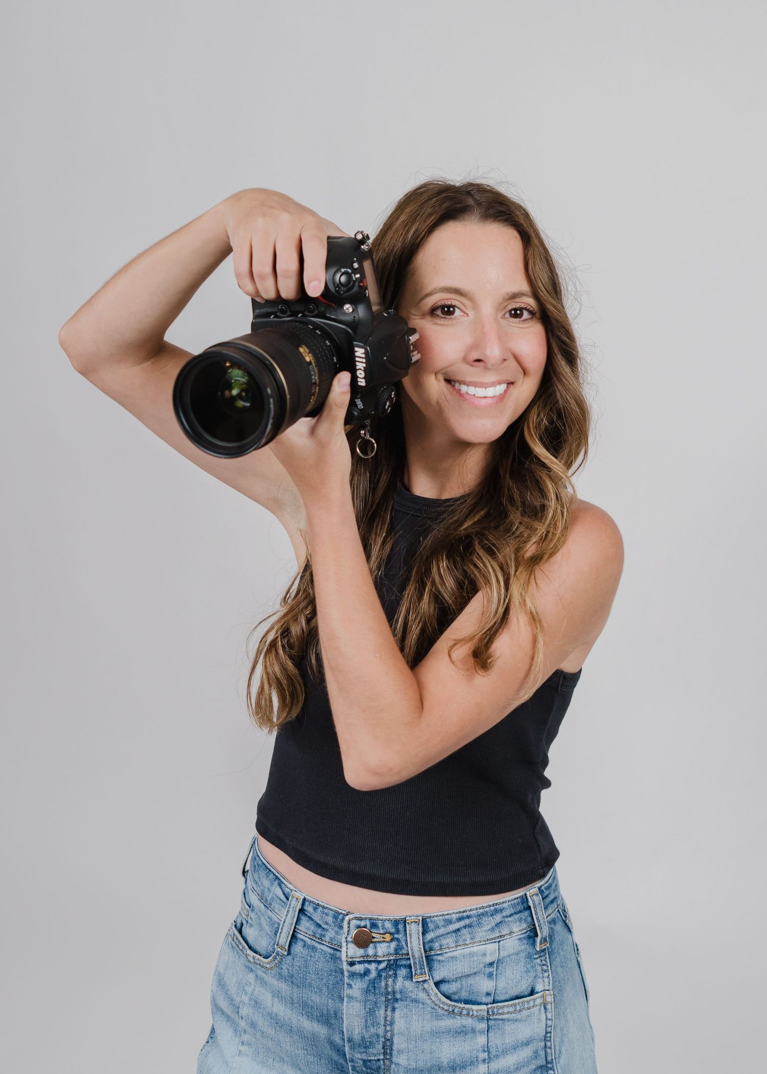 Professional photographer holding a DSLR camera while wearing a black crop top and jeans against a light gray background.