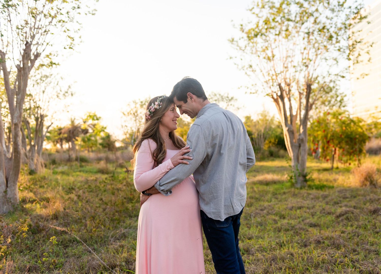 A couple embraces in a sunlit wooded area during sunset while wearing pink and grey casual attire.