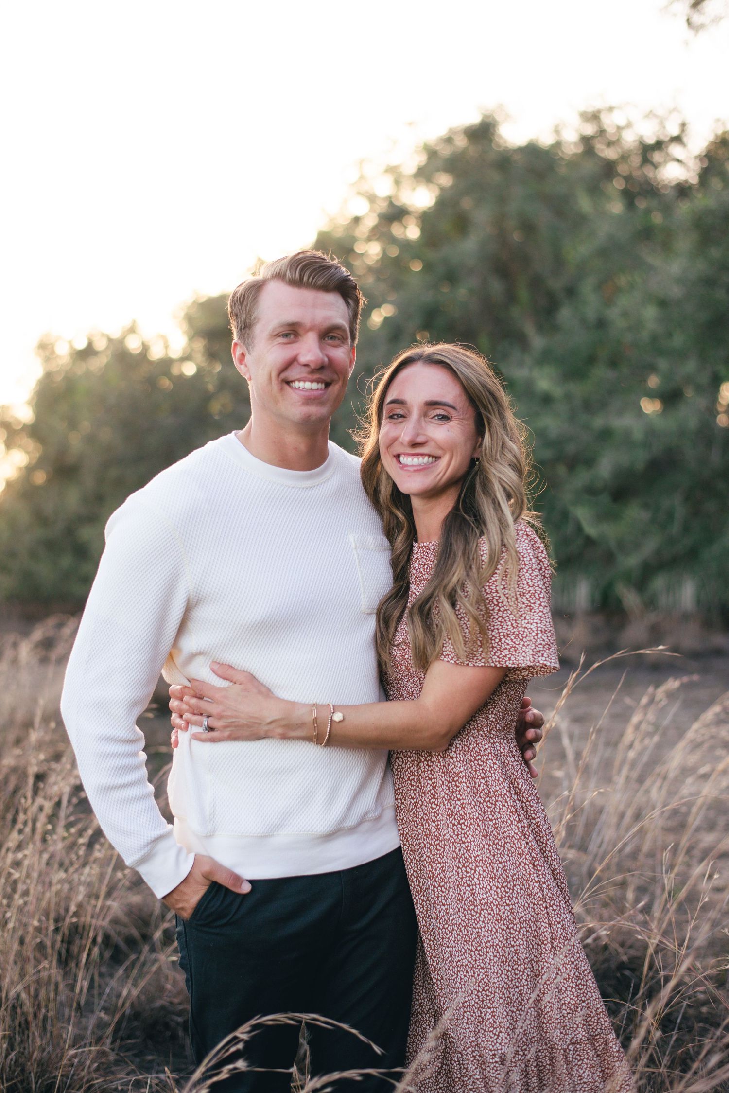 A couple in love poses together in a field at sunset, wearing a white sweater and floral pink dress.