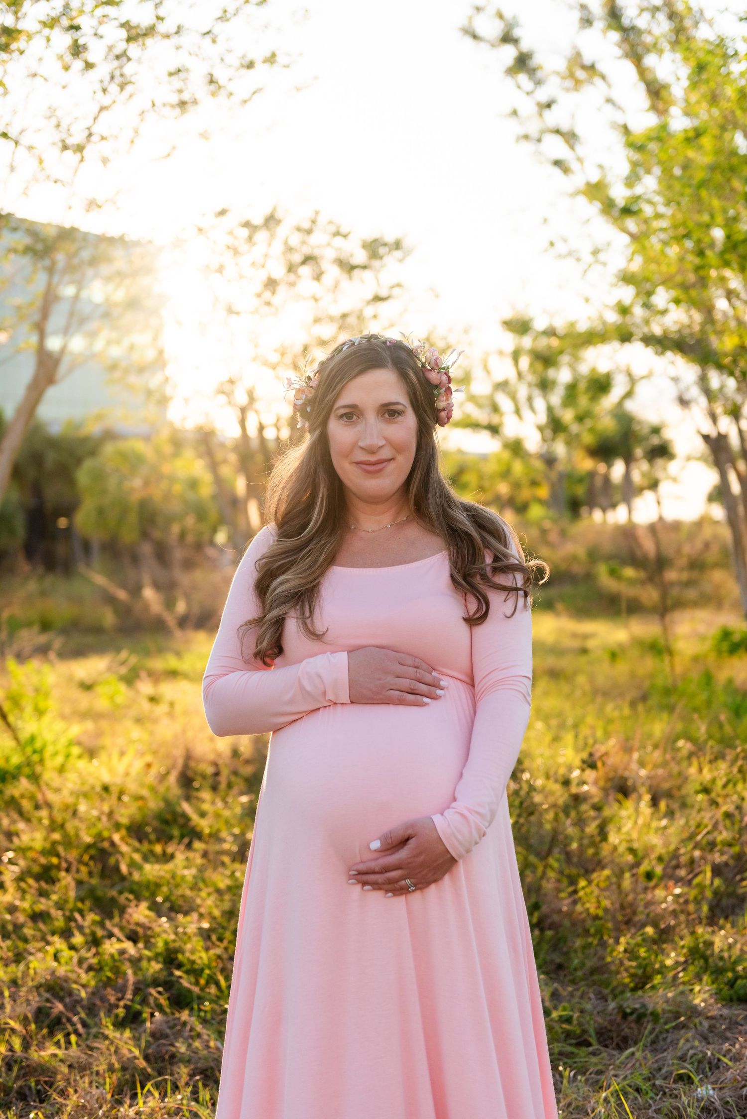 A person in a pink long-sleeved dress stands in a sunlit field during golden hour, cradling their stomach tenderly.