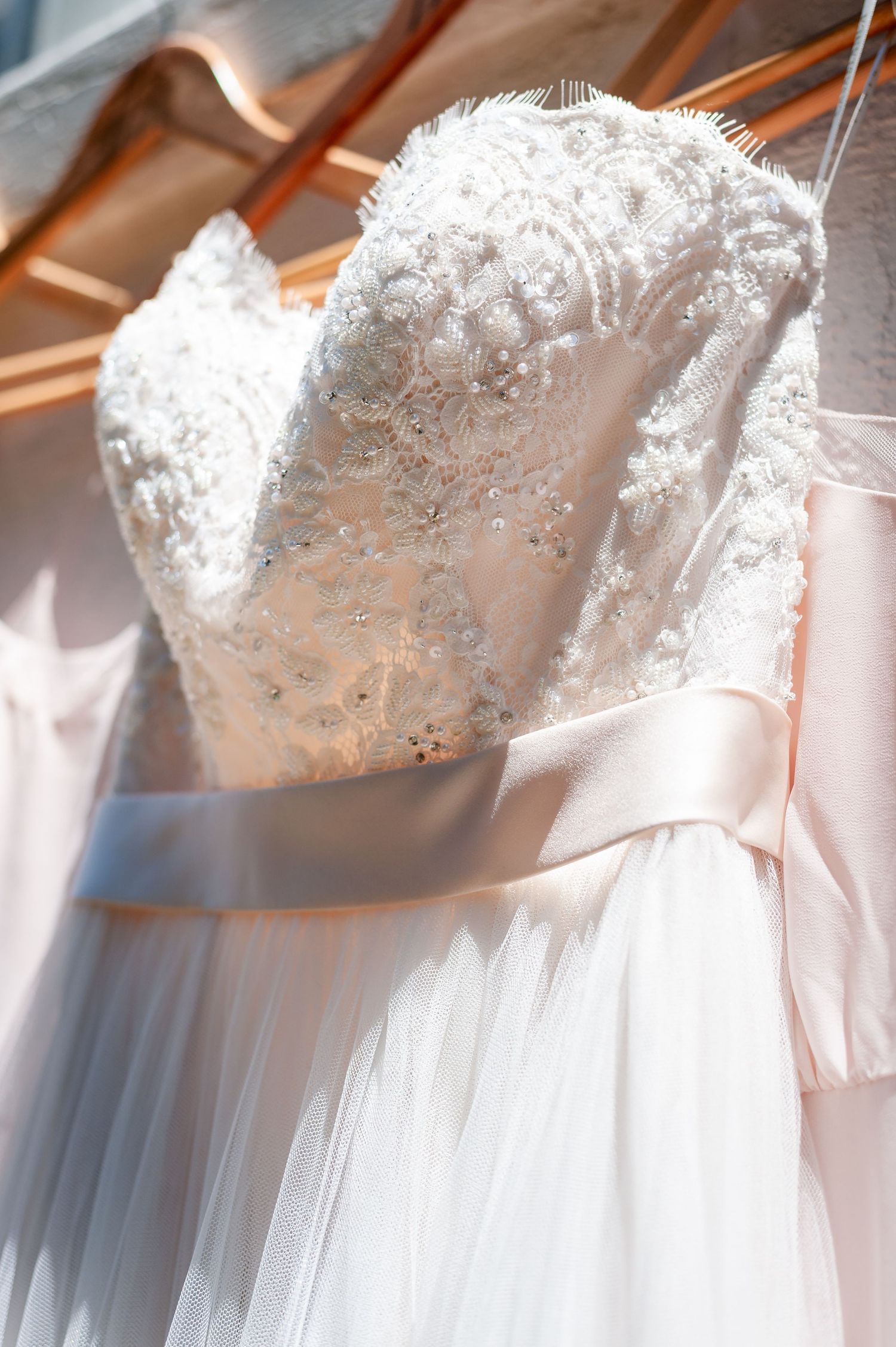 A white lace wedding dress with delicate beading and a satin ribbon sash hangs gracefully.