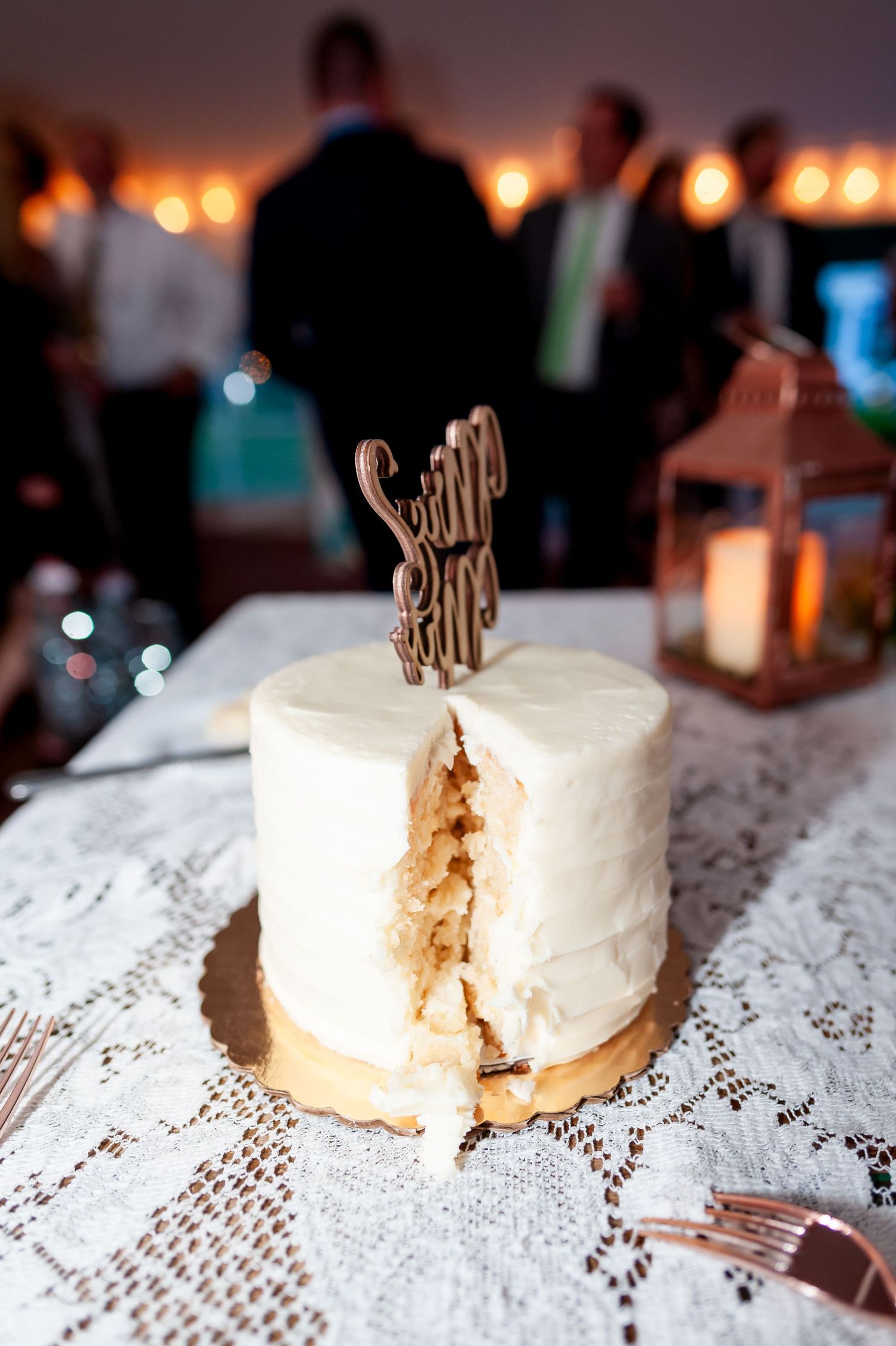 White wedding cake with rustic topper showing slice on lace tablecloth with lantern decoration.