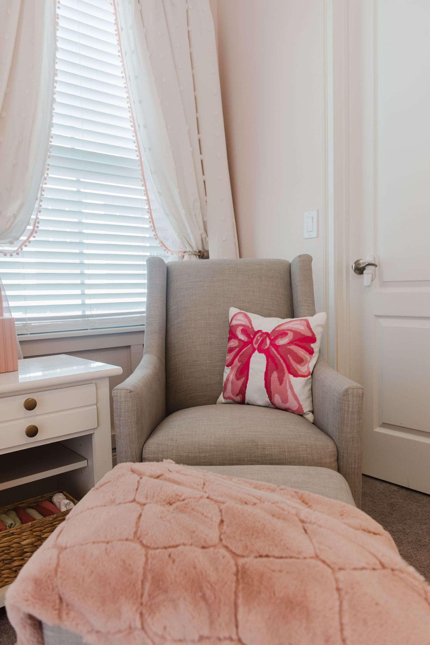 Pink and gray nursery corner with plush bedding, curtains, and a decorative bow pillow.