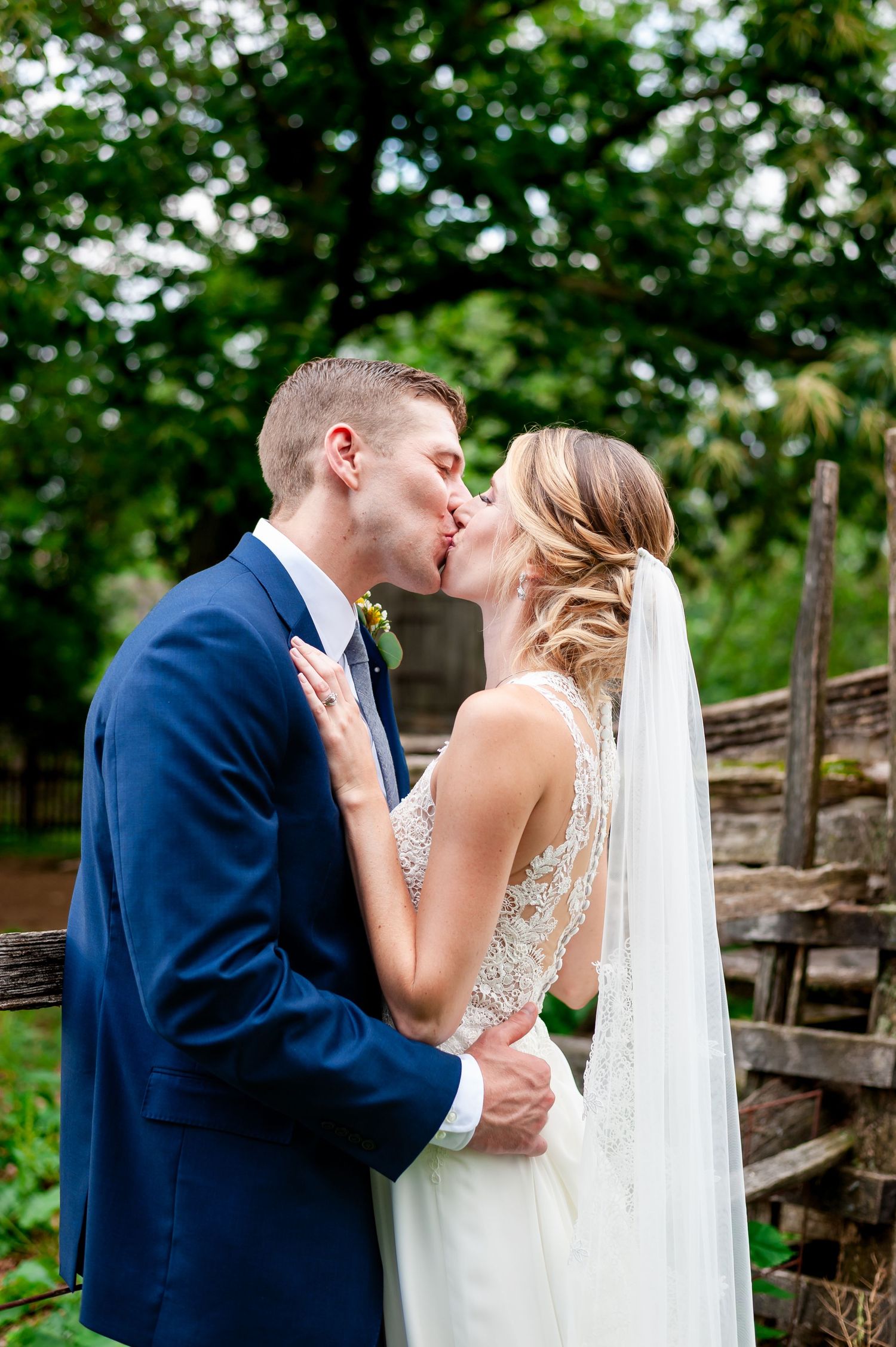 A romantic wedding kiss captured in front of lush greenery with the bride wearing a detailed lace gown and veil.