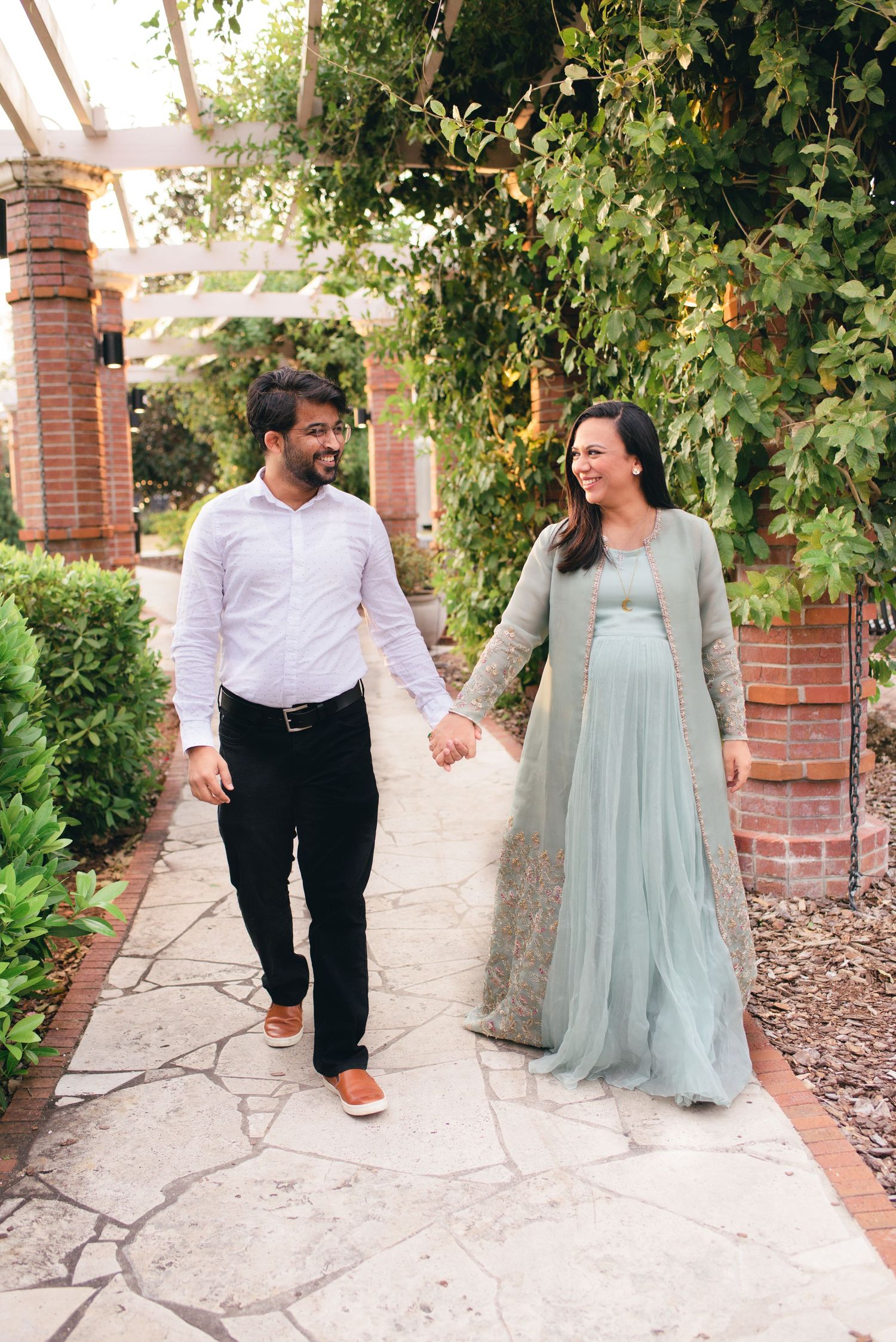 Two individuals in formal attire walk hand in hand along a stone path surrounded by lush garden and brick columns.
