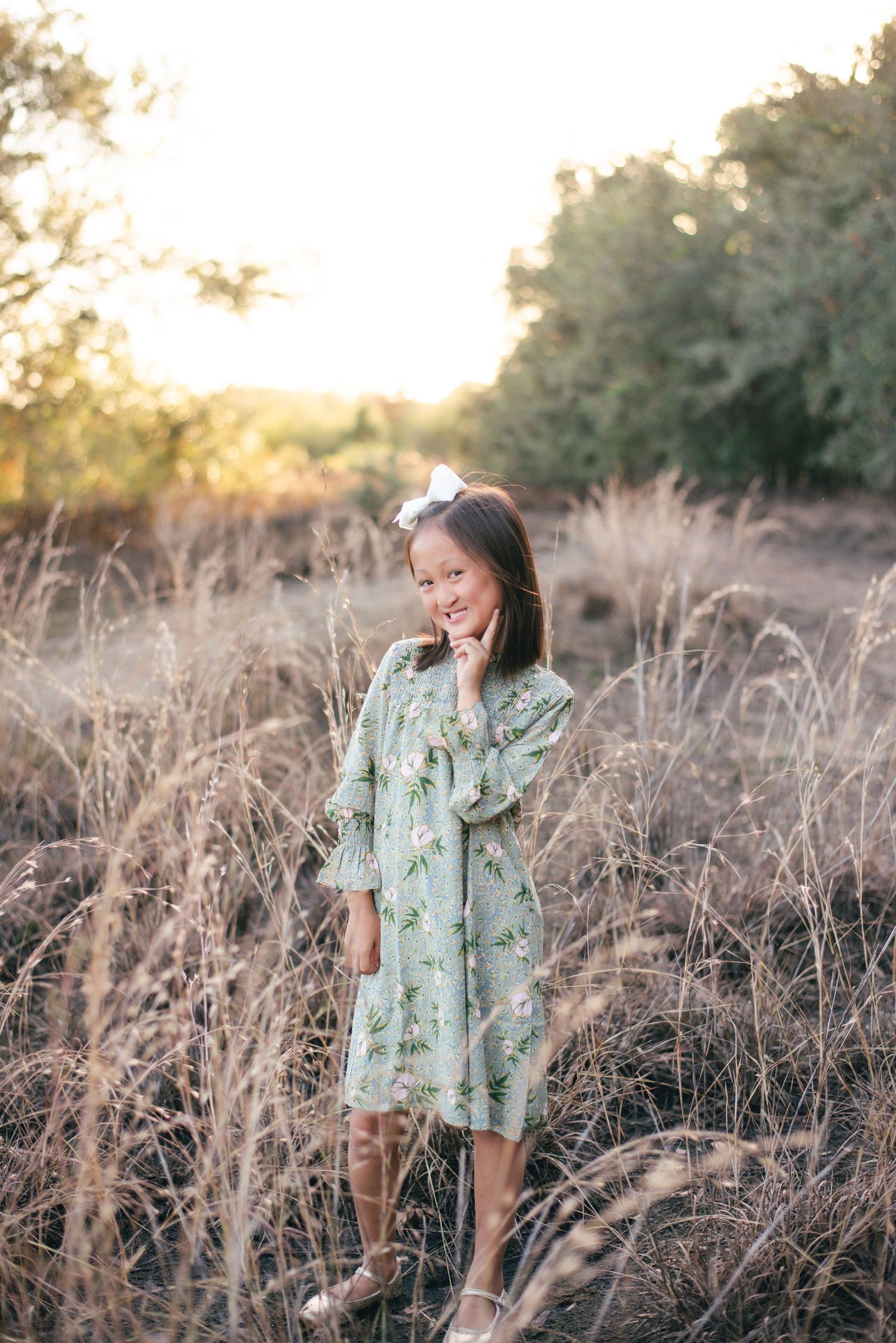 A figure stands in a mint green floral dress among tall dried grass during golden hour sunset.