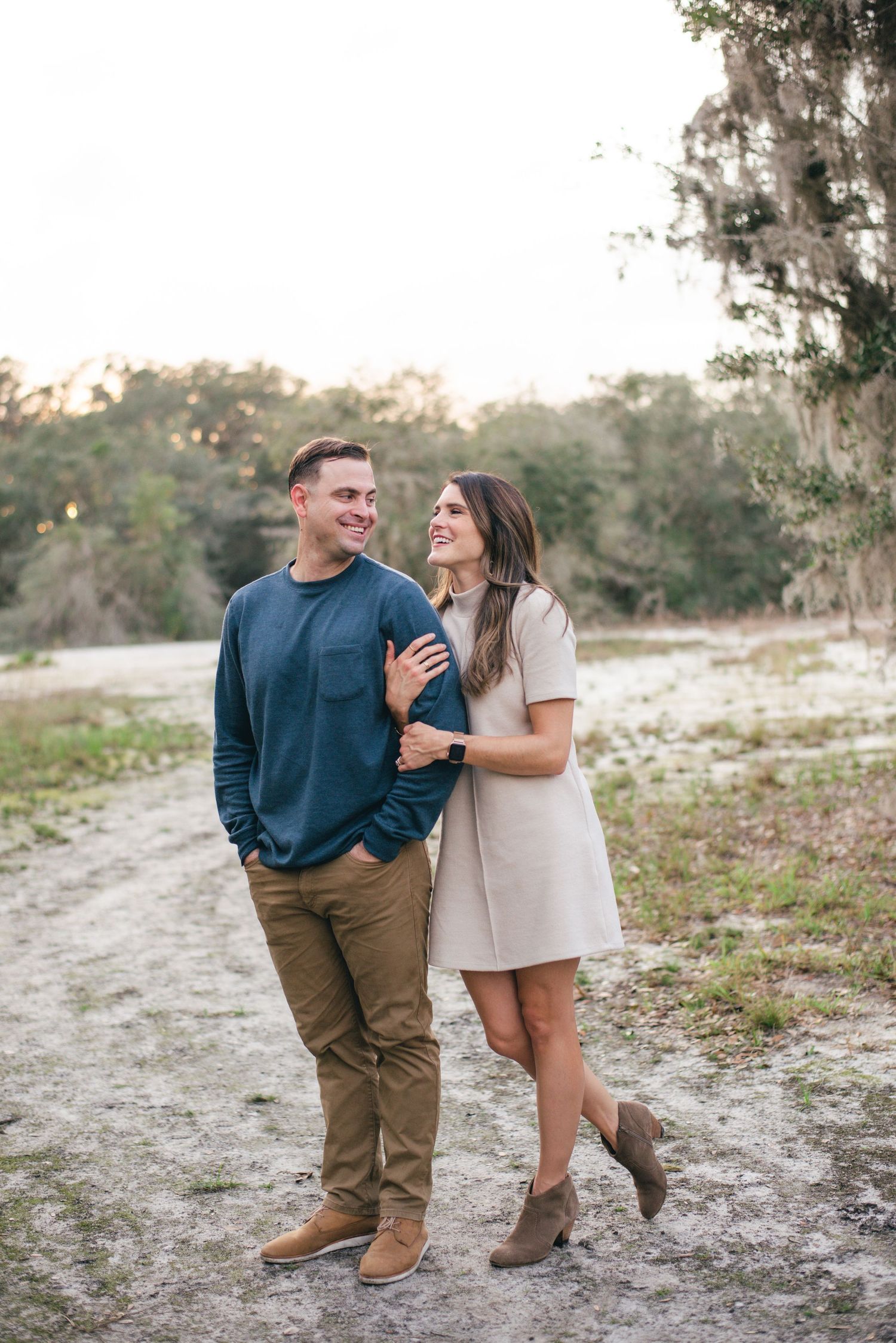 A couple shares a joyful moment together while standing on a sandy path surrounded by trees at sunset.