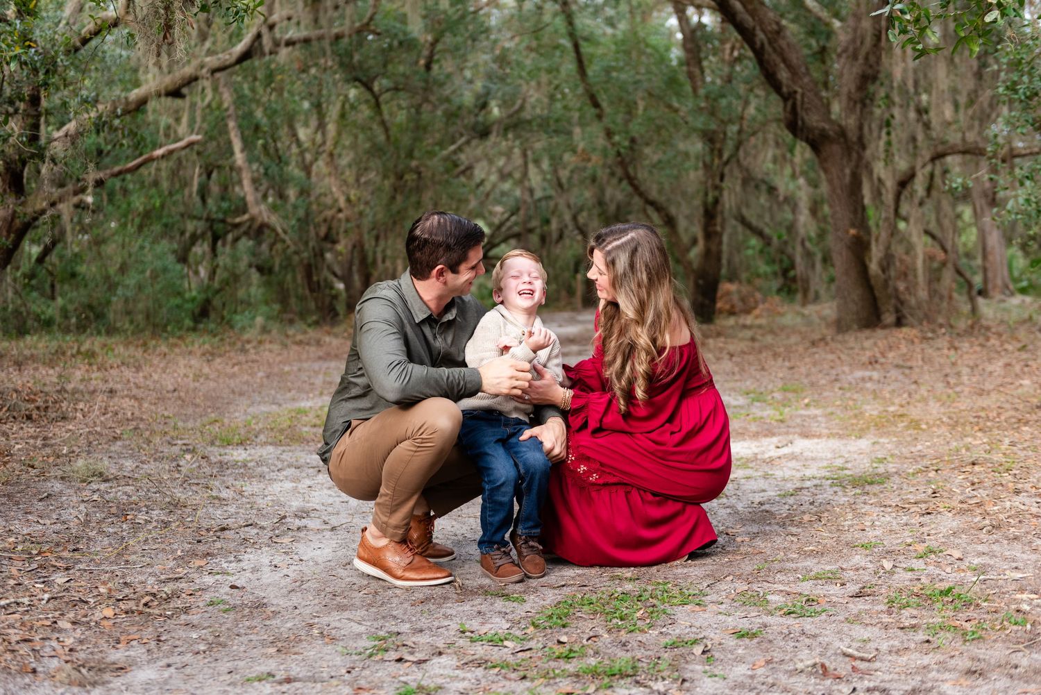 Two people in red and navy outfits sit together on a sandy path in a wooded outdoor setting.