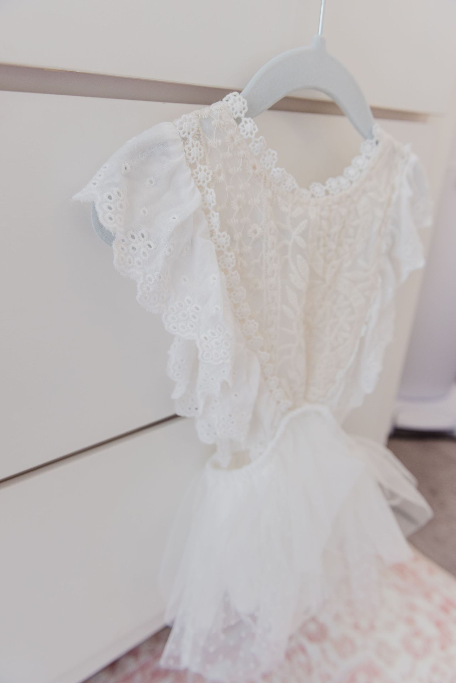 Delicate white lace baby dress with ruffled sleeves hanging on a white hanger.