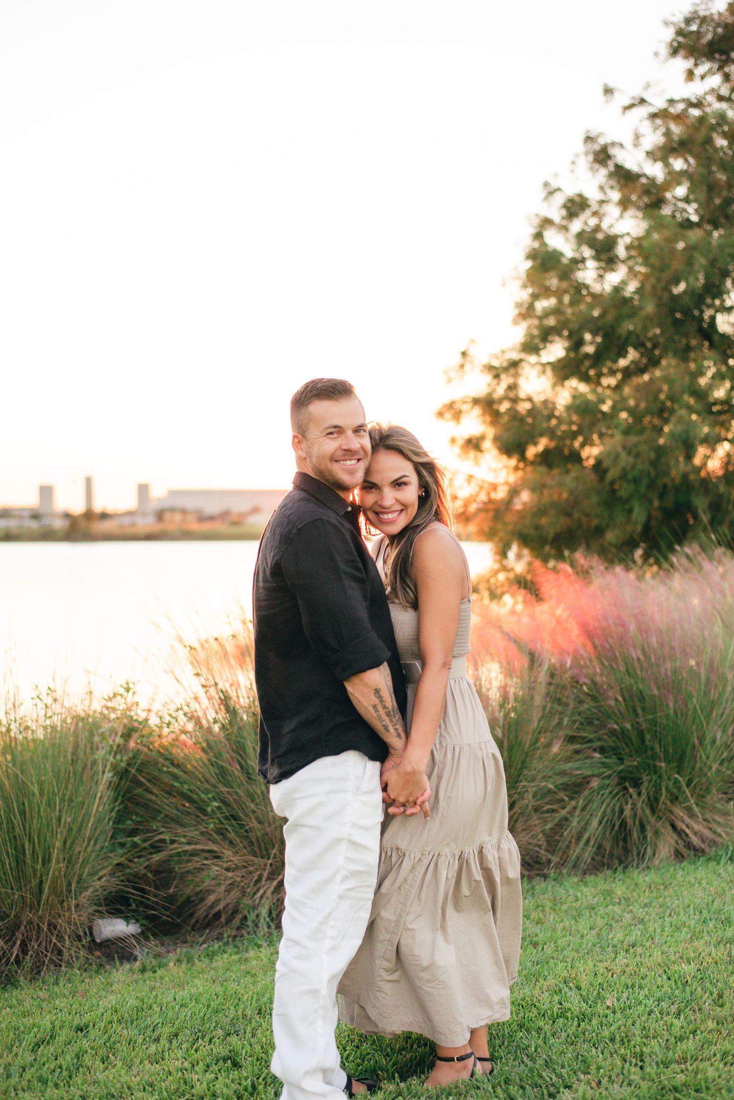 A couple embraces on grassy riverbank at sunset with ornamental grasses and trees in the background.