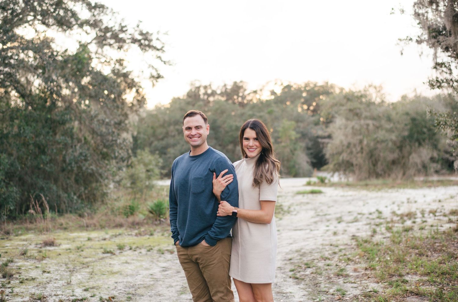 Couple stands close together in casual attire on a sandy path during golden hour with trees in background.