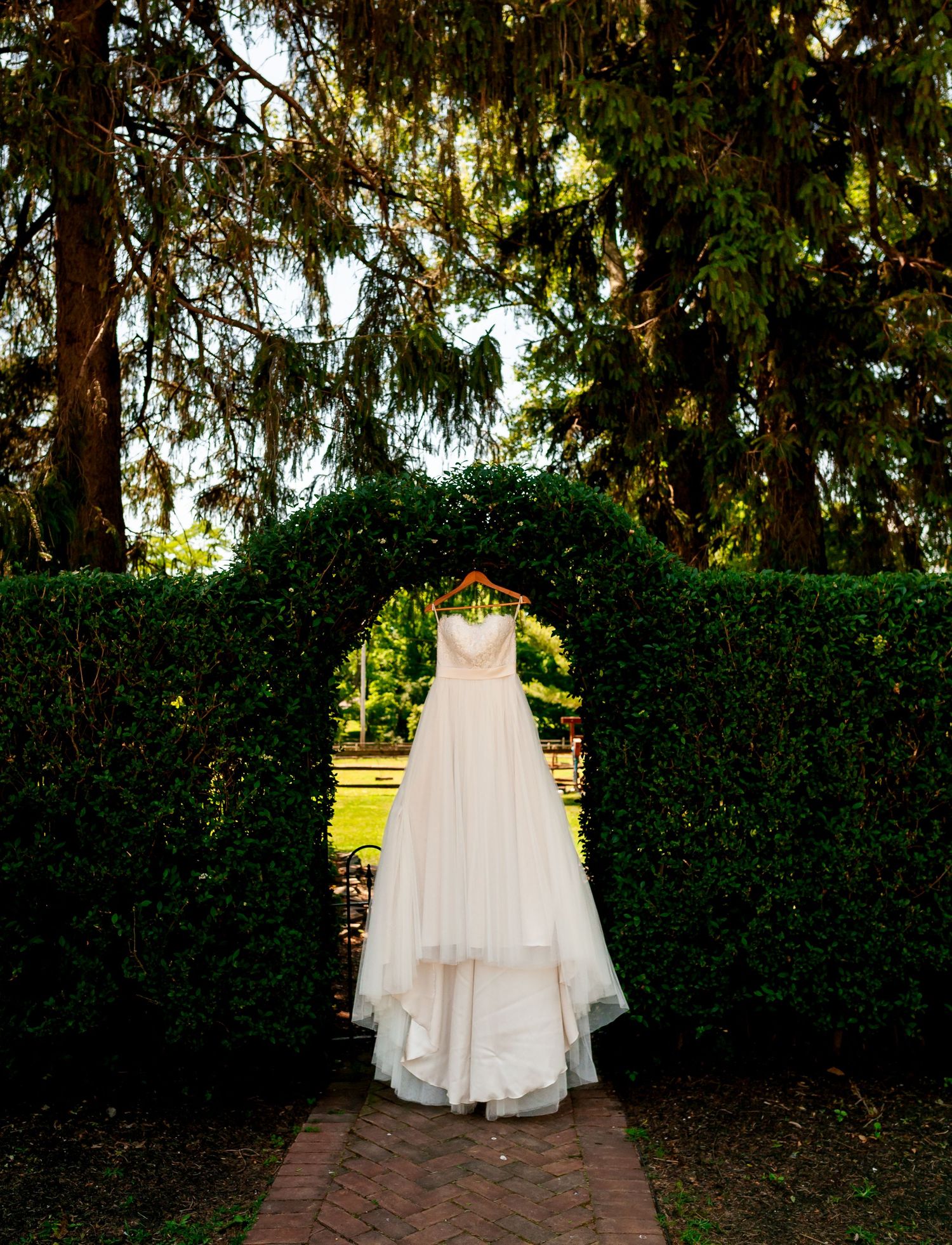 Elegant white wedding gown displayed in garden archway with sunlight filtering through trees.