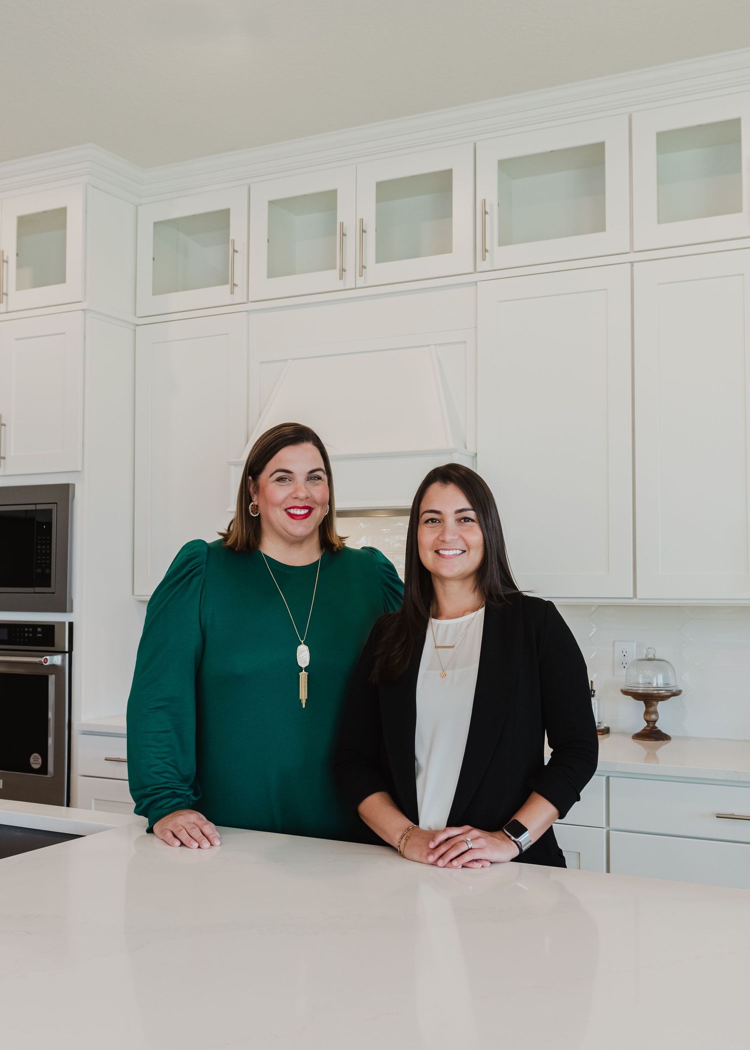 Two professionals in business attire posing together in a modern white kitchen.