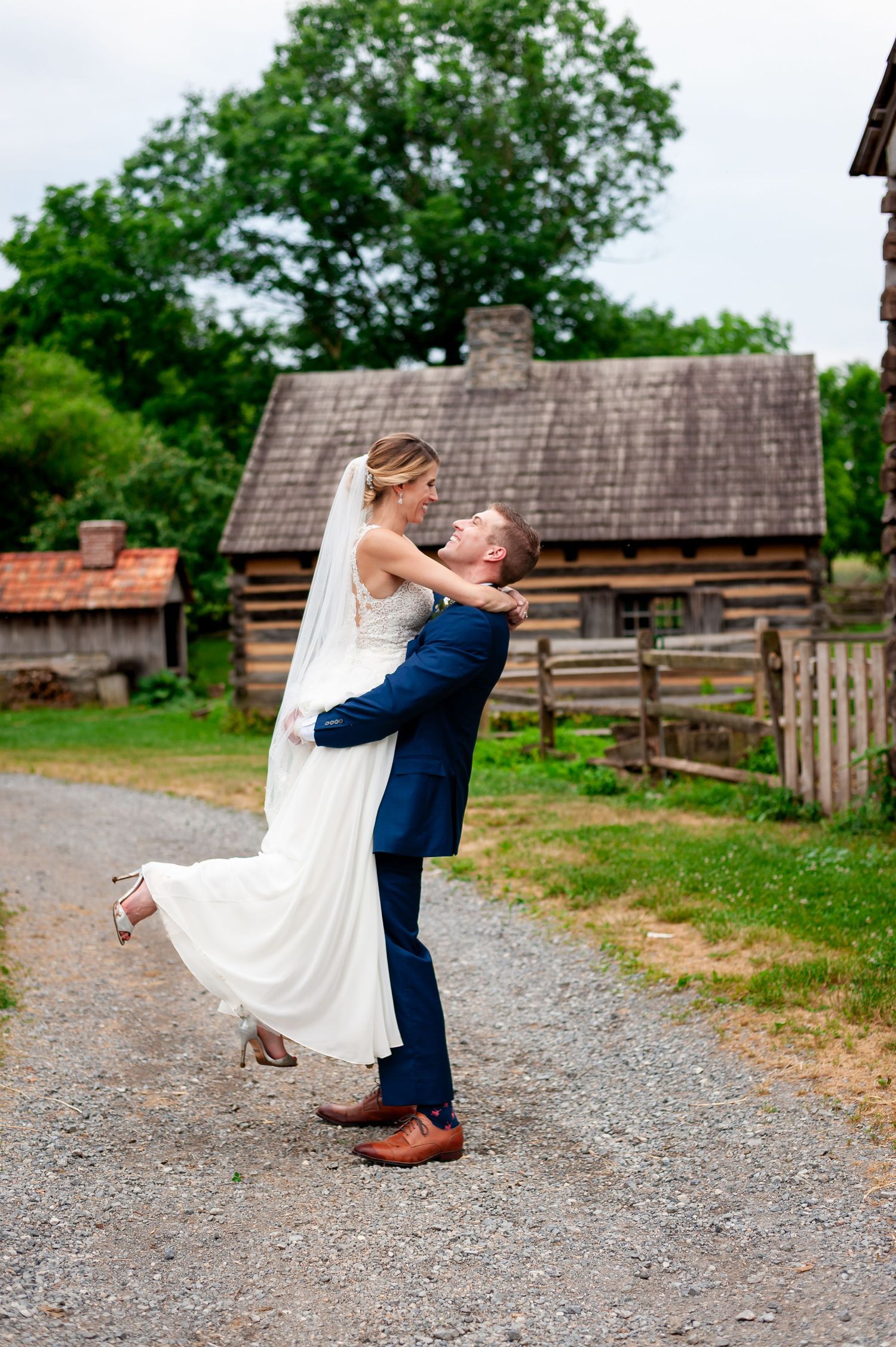 Playful wedding couple embrace on a country path with historic log buildings behind them.