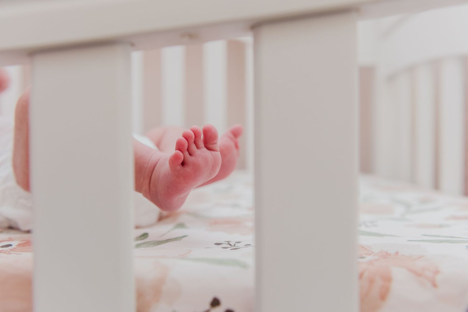 Close-up view of tiny feet through white crib slats with floral bedding.