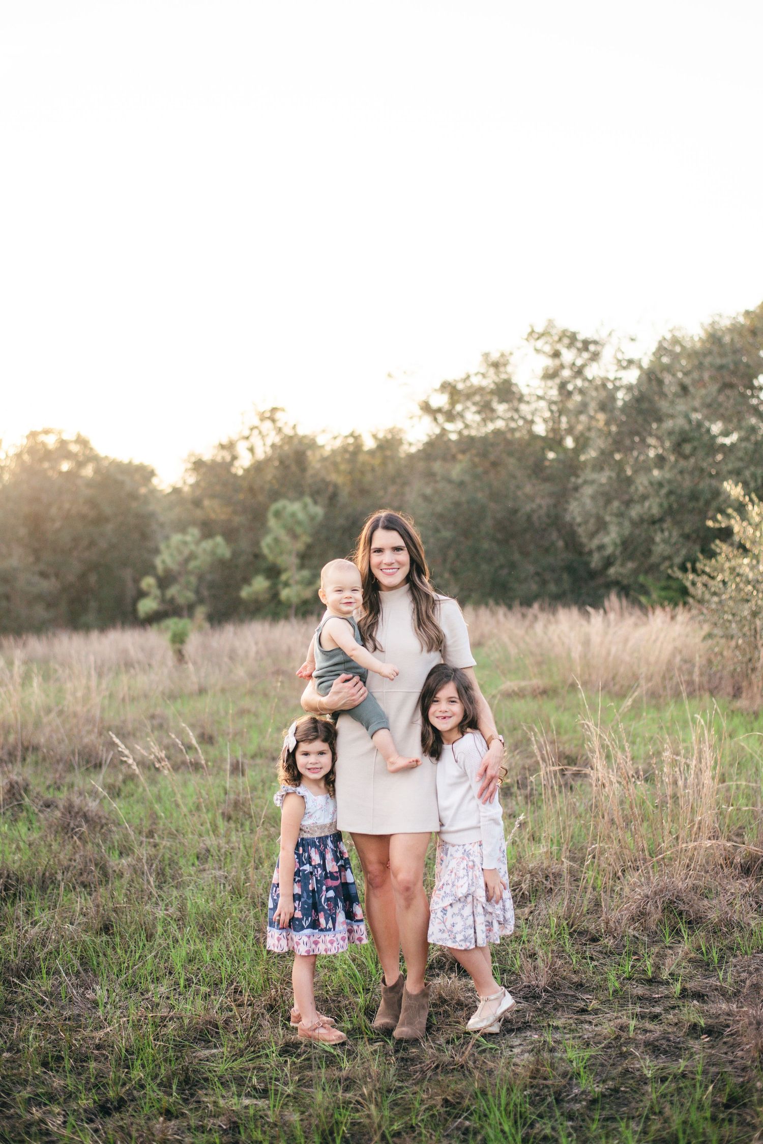 A parent and children pose together in a rustic field setting at golden hour with tall grass and trees in the background.