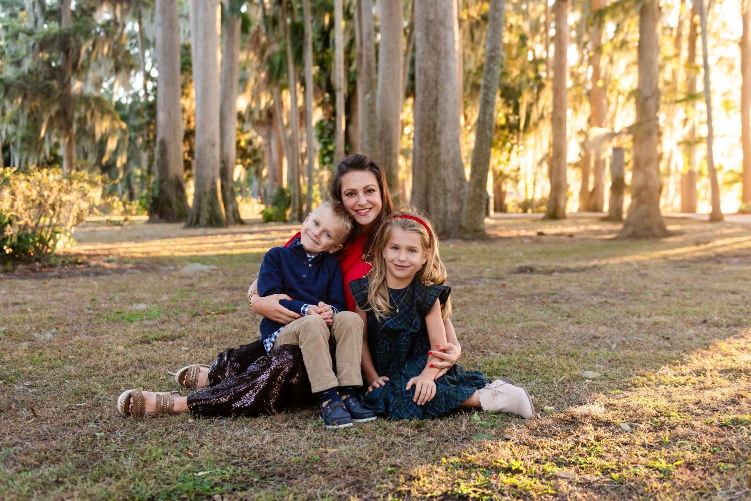 Three people sitting together on fallen leaves in a forest during golden hour sunlight filtering through tall trees.