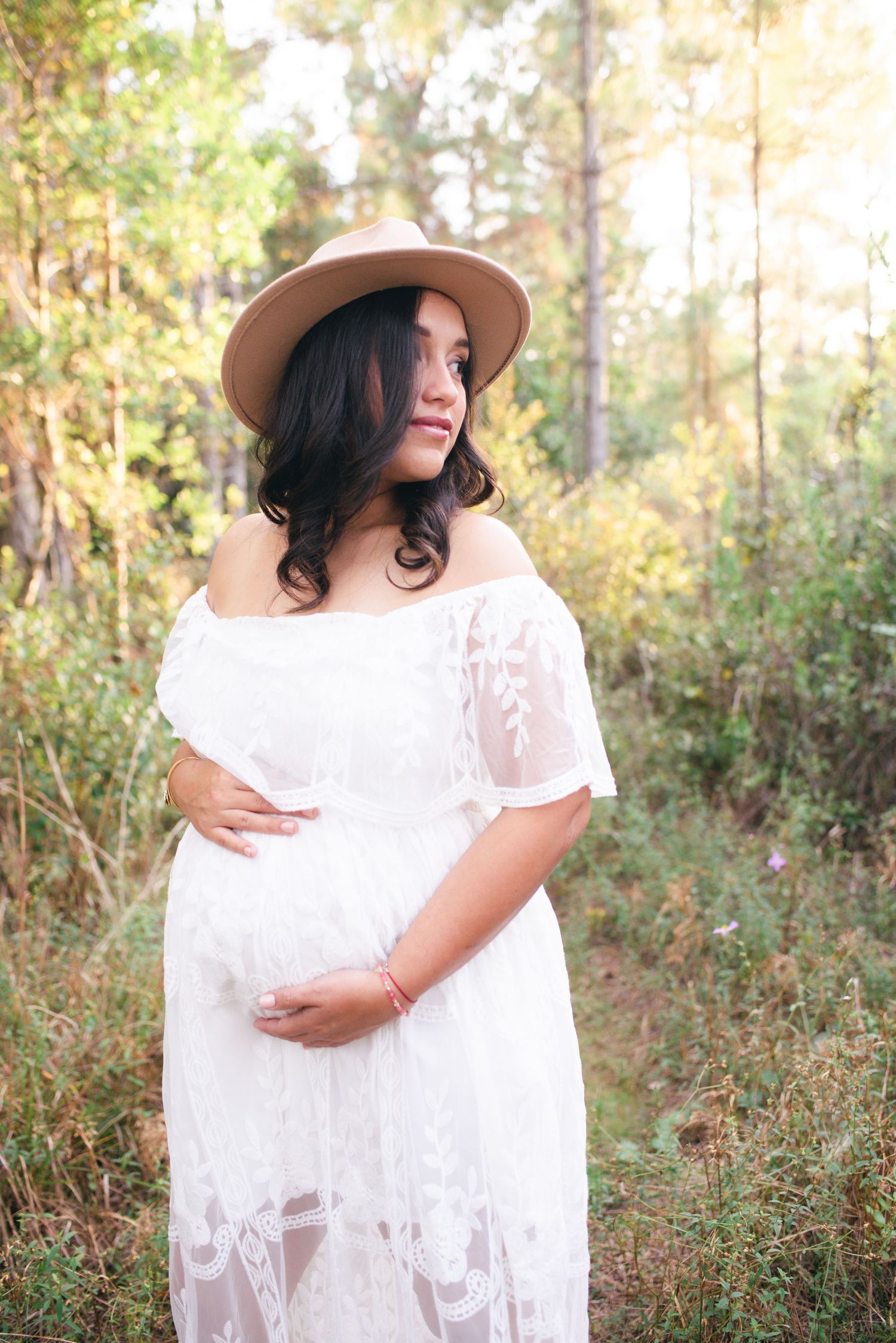 Woman in white lace dress and sun hat poses gracefully in a sunlit forest setting during an outdoor photo session.