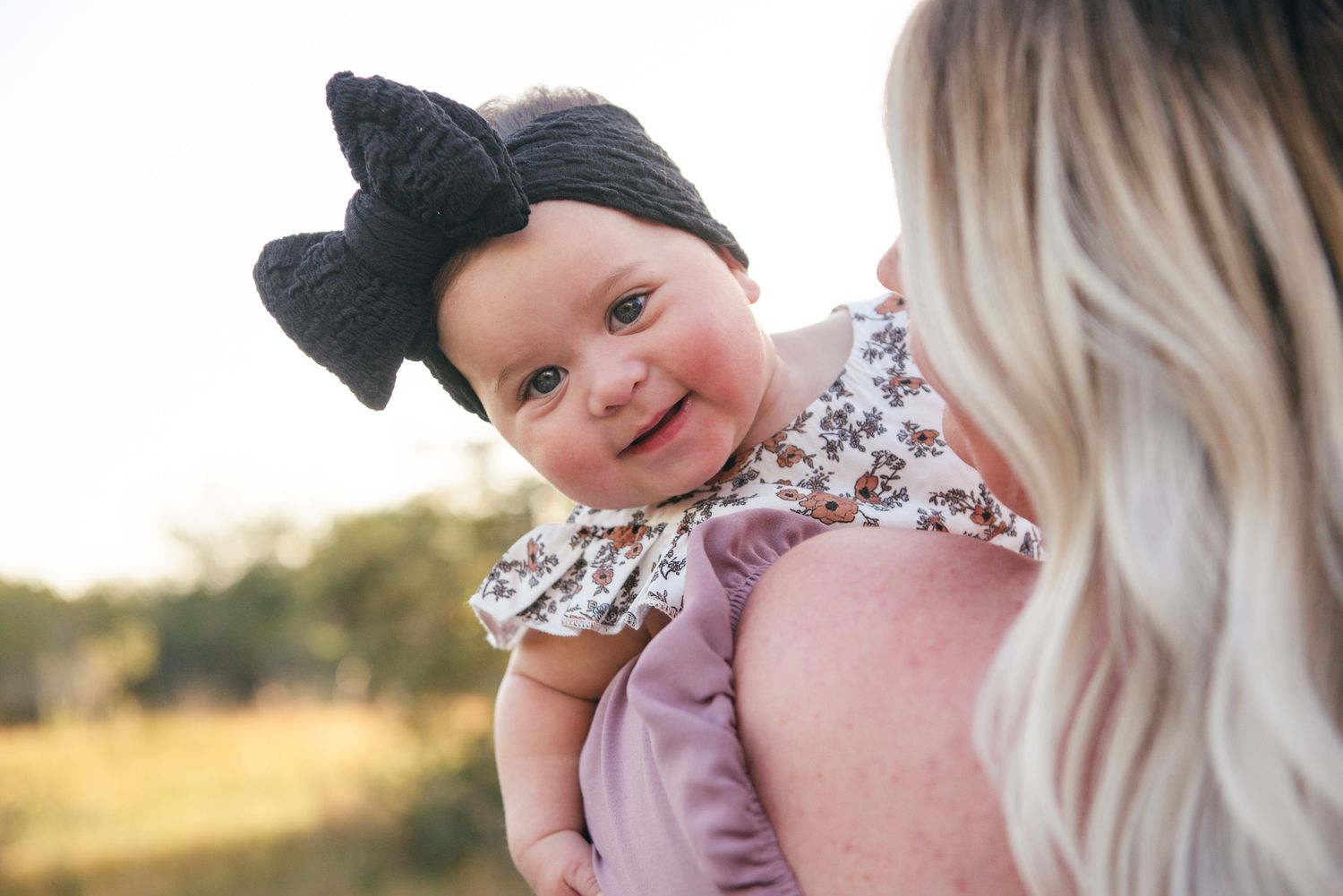 Smiling baby wearing black bow headband and floral outfit in outdoor portrait.