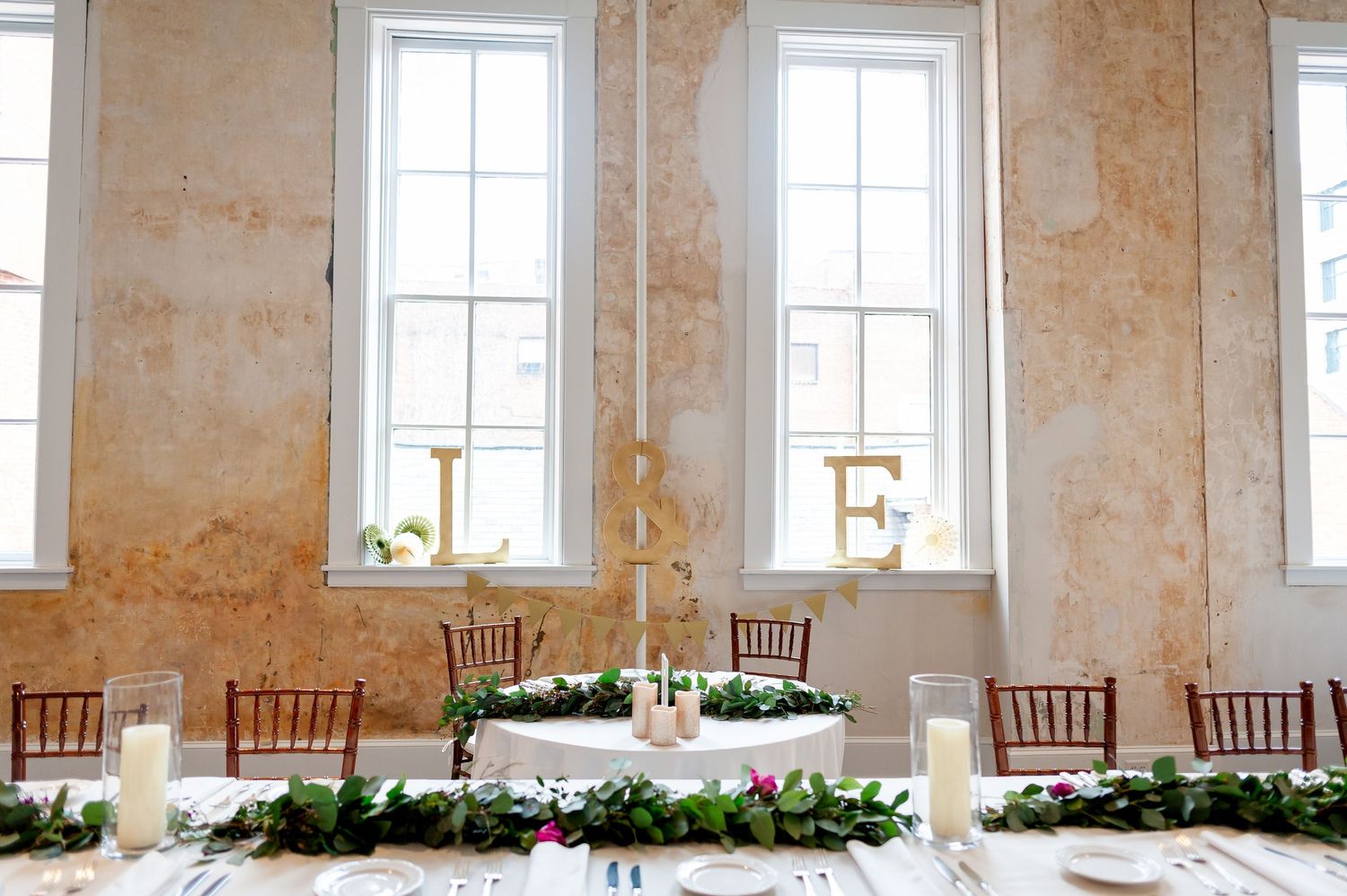 Elegant wedding reception table setup with tall windows and rustic walls.