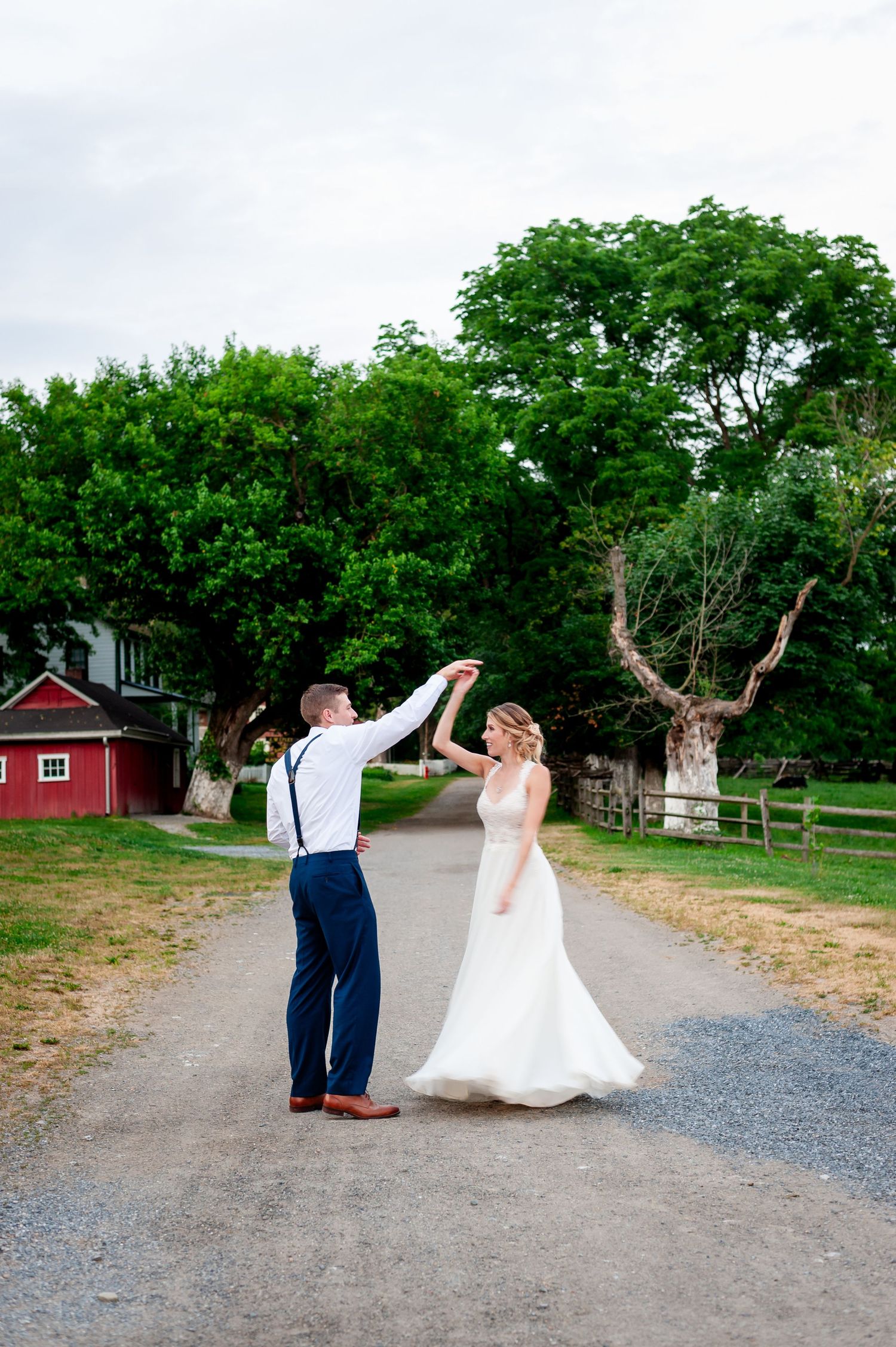Newlyweds dance joyfully on a path with a red barn and trees in the background.
