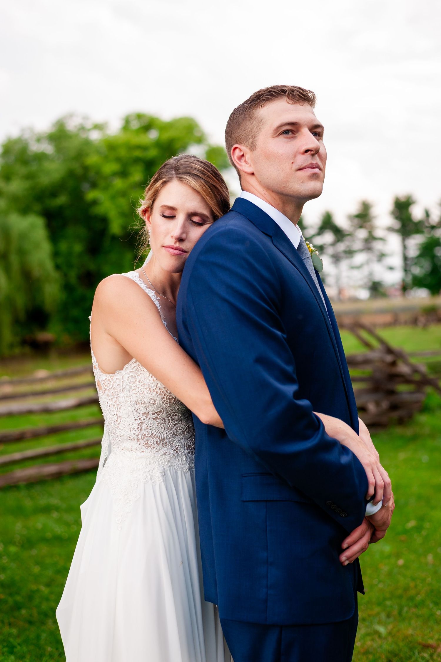 Elegant couple in navy suit and white dress pose against verdant countryside backdrop.