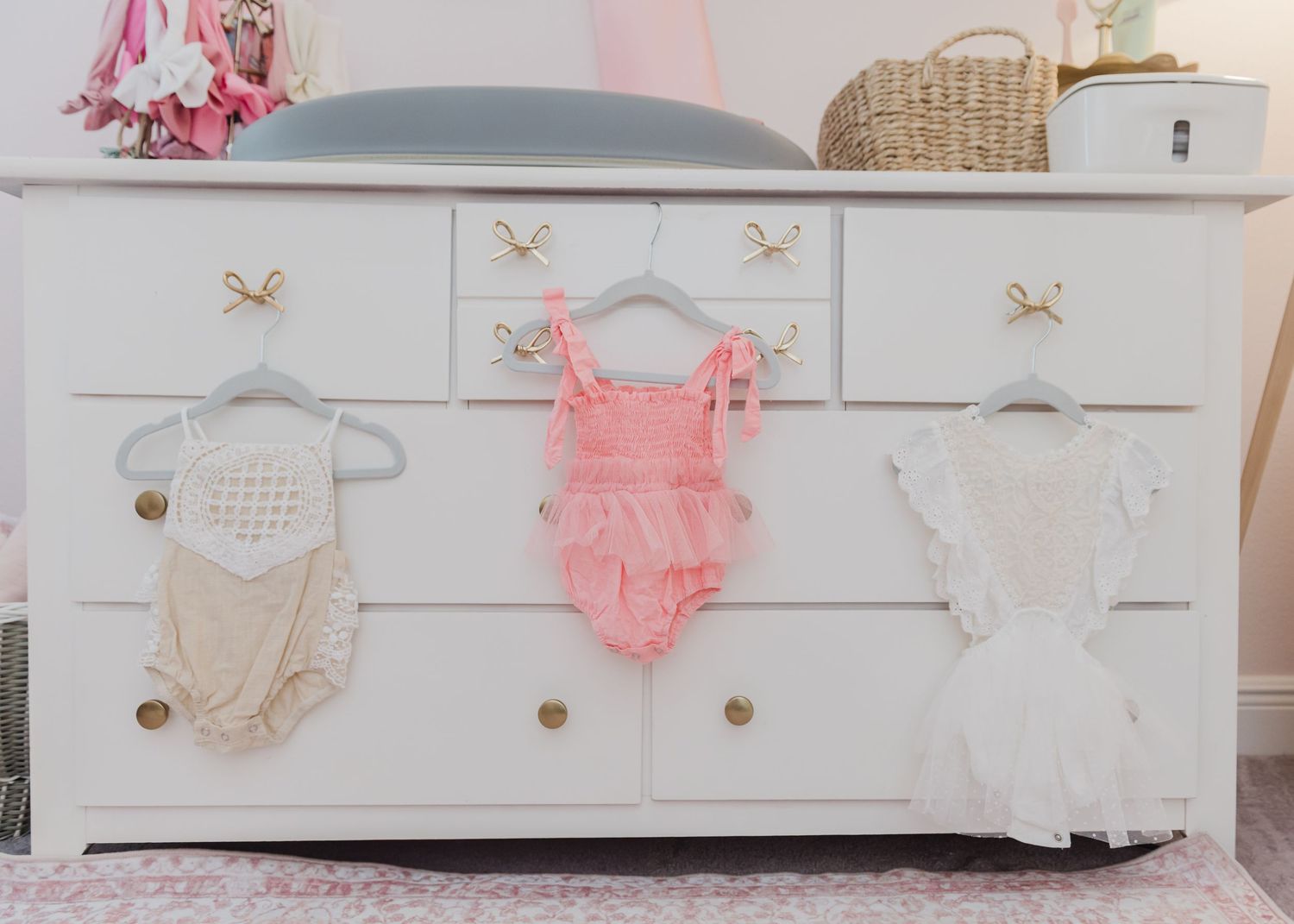 Pink and white baby clothes displayed on hangers against a white dresser in a nursery.