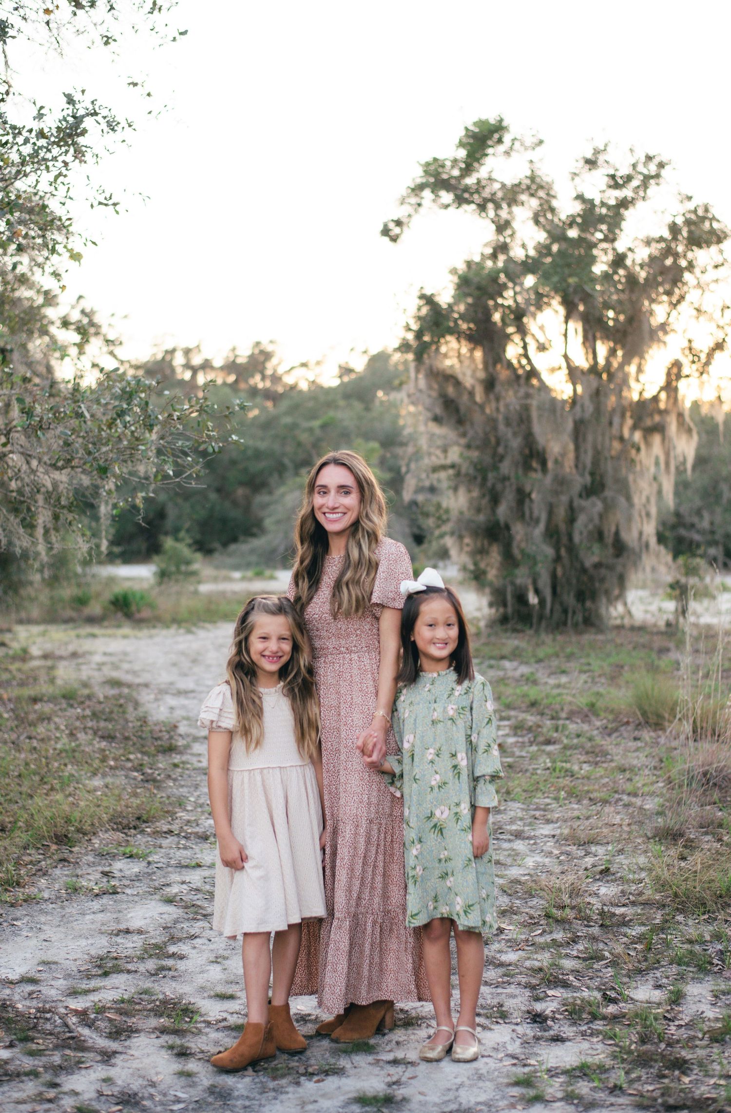 Three people in flowing maxi dresses stand together on sandy ground at sunset with Spanish moss trees in background.