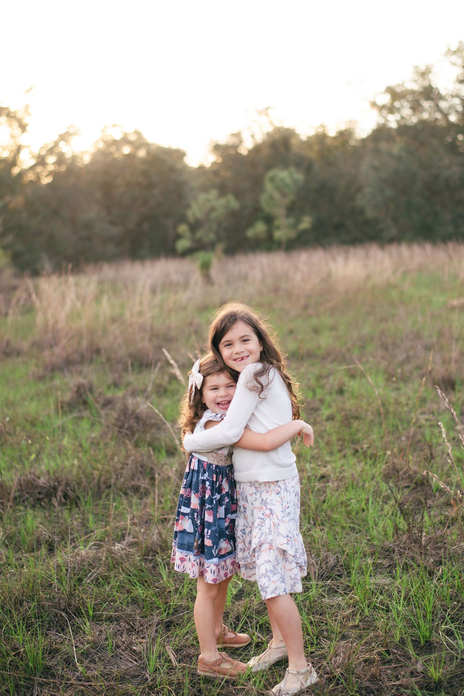 Two young girls in patterned dresses share a sweet embrace while standing in a sunlit meadow at sunset.