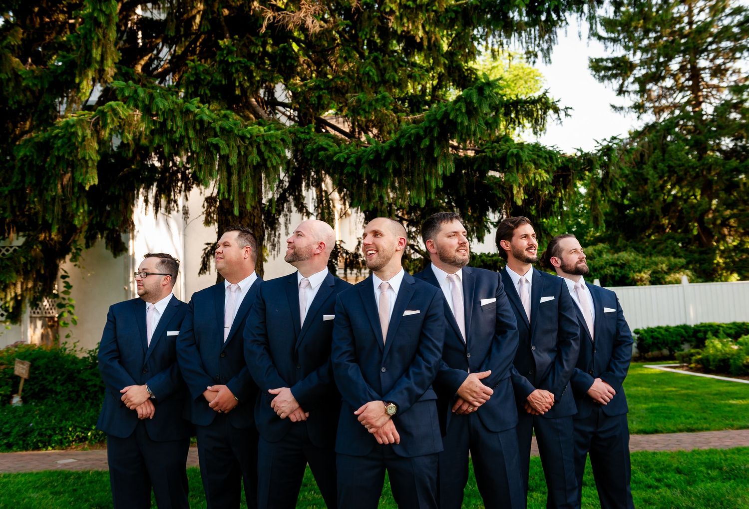 Groomsmen in matching navy suits and pink ties stand in line outdoors.