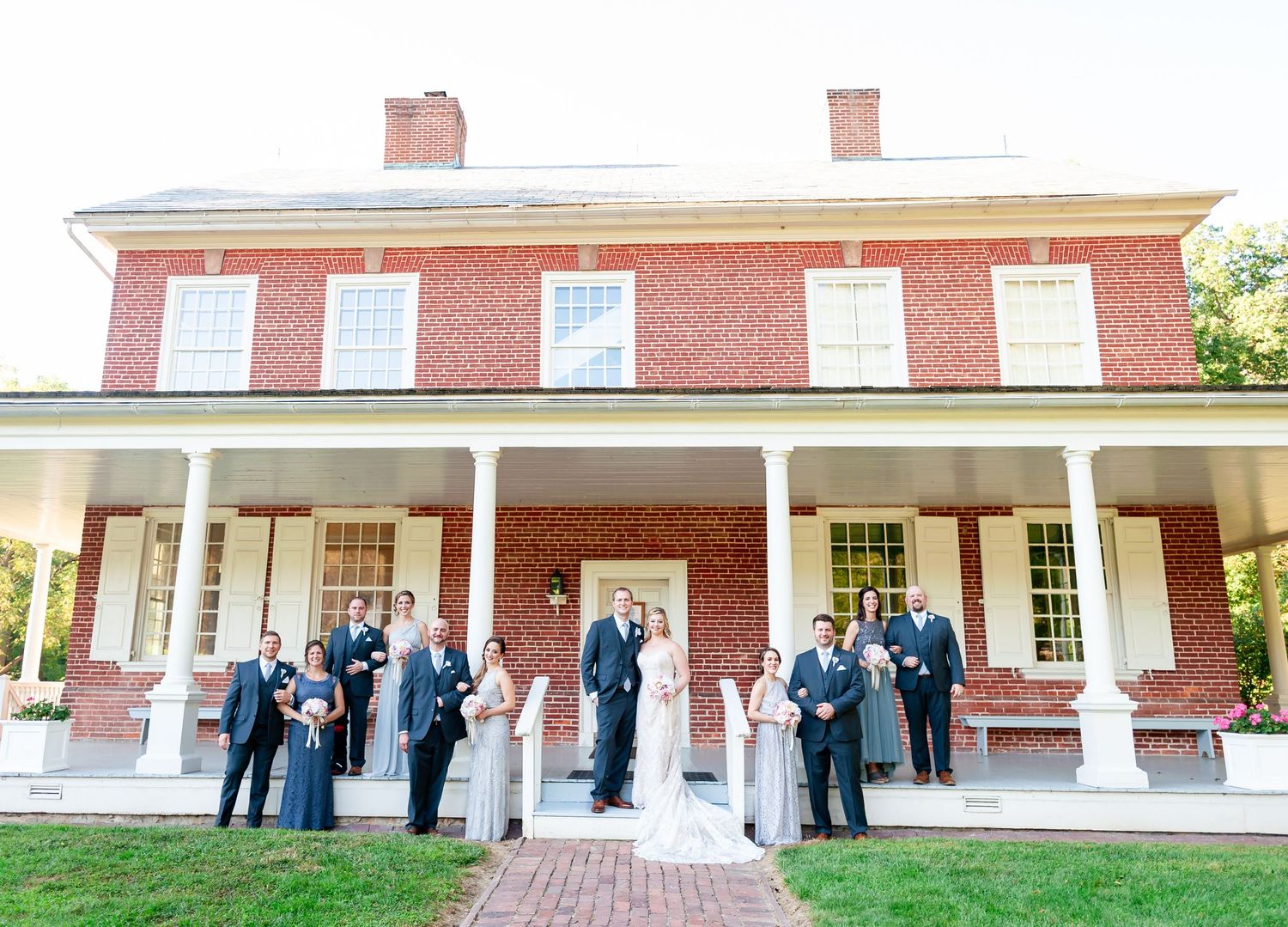 Wedding party poses along brick building's white columned porch in formal attire on sunny day.