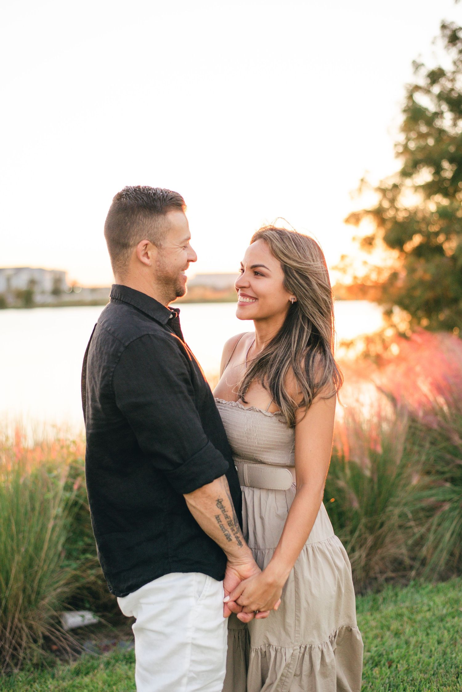 A couple holding hands by a lake at sunset, with tall grass and golden light creating a romantic outdoor setting.