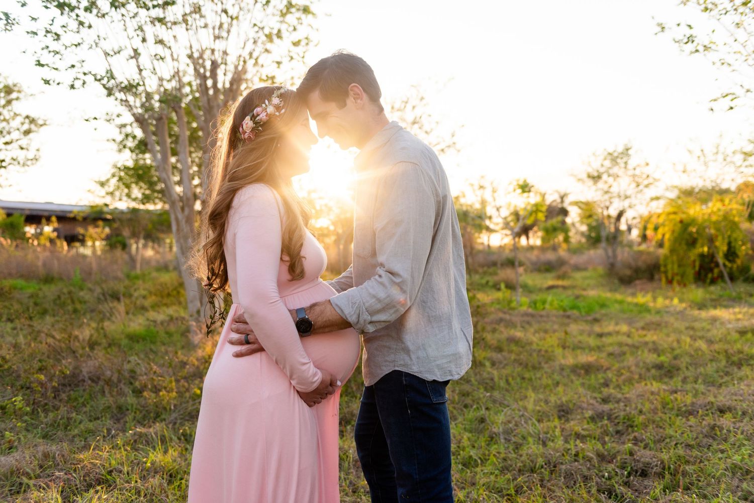 Couple embraces in a meadow during golden hour as sunlight streams through trees creating a romantic silhouette.