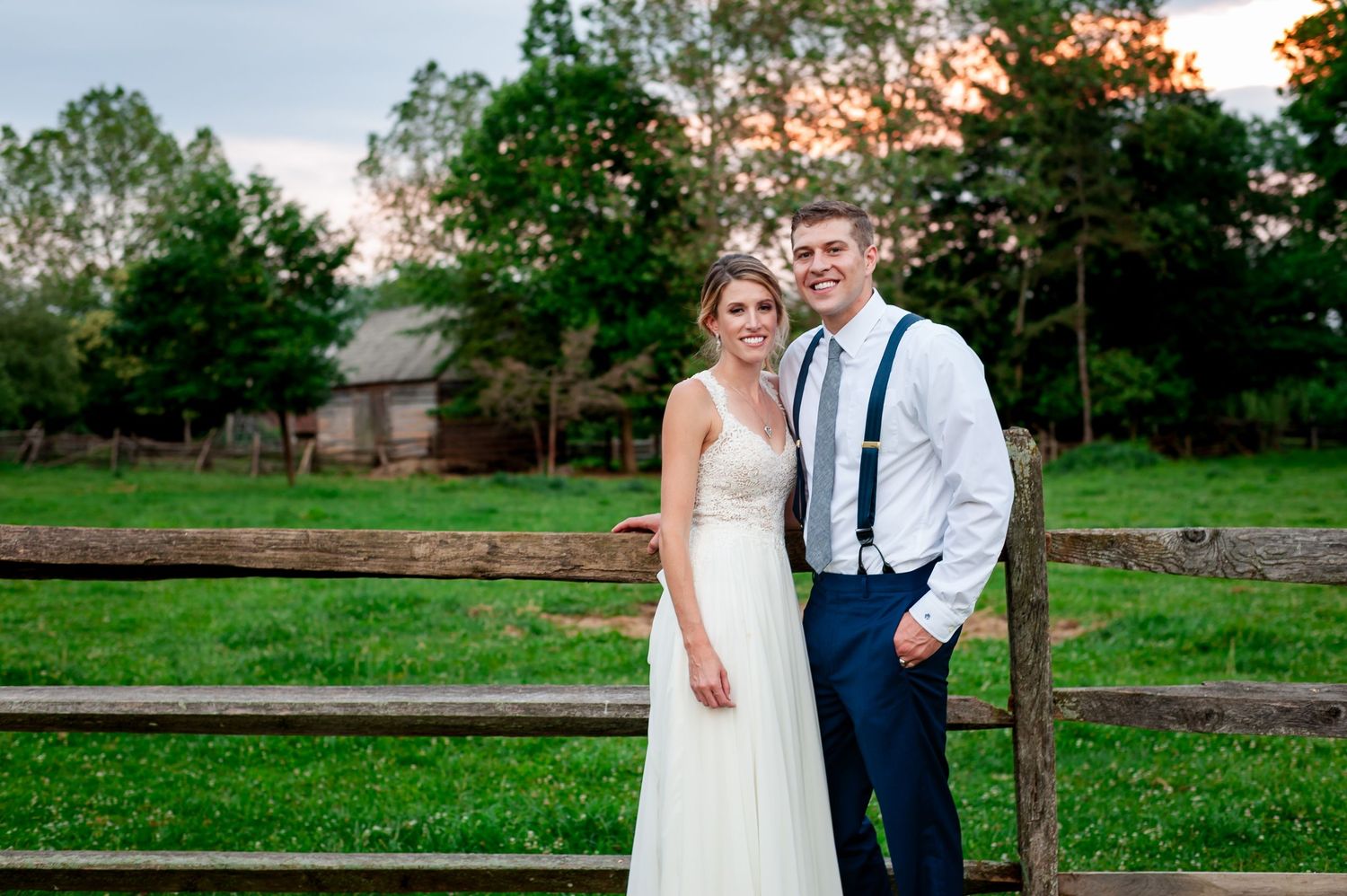 Sunset wedding portrait with couple standing by wooden fence in rustic farm setting.