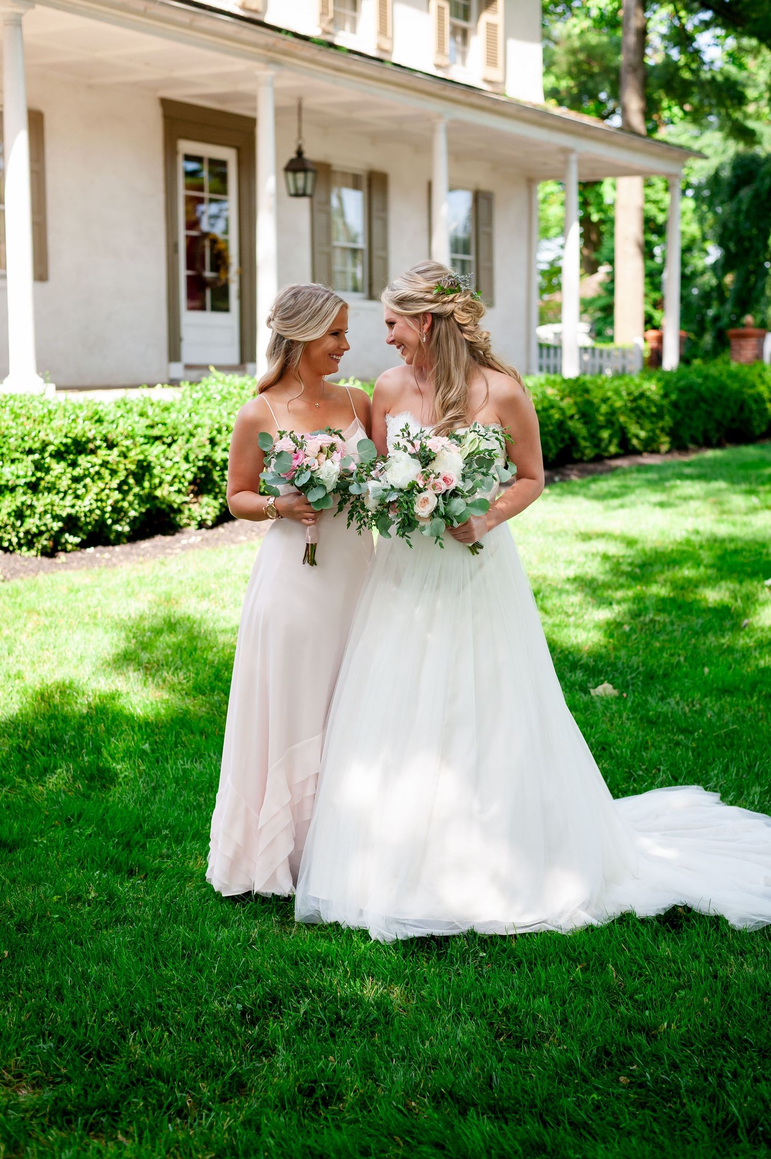 Two people in elegant dresses holding bouquets on a manicured lawn in front of a classic house.