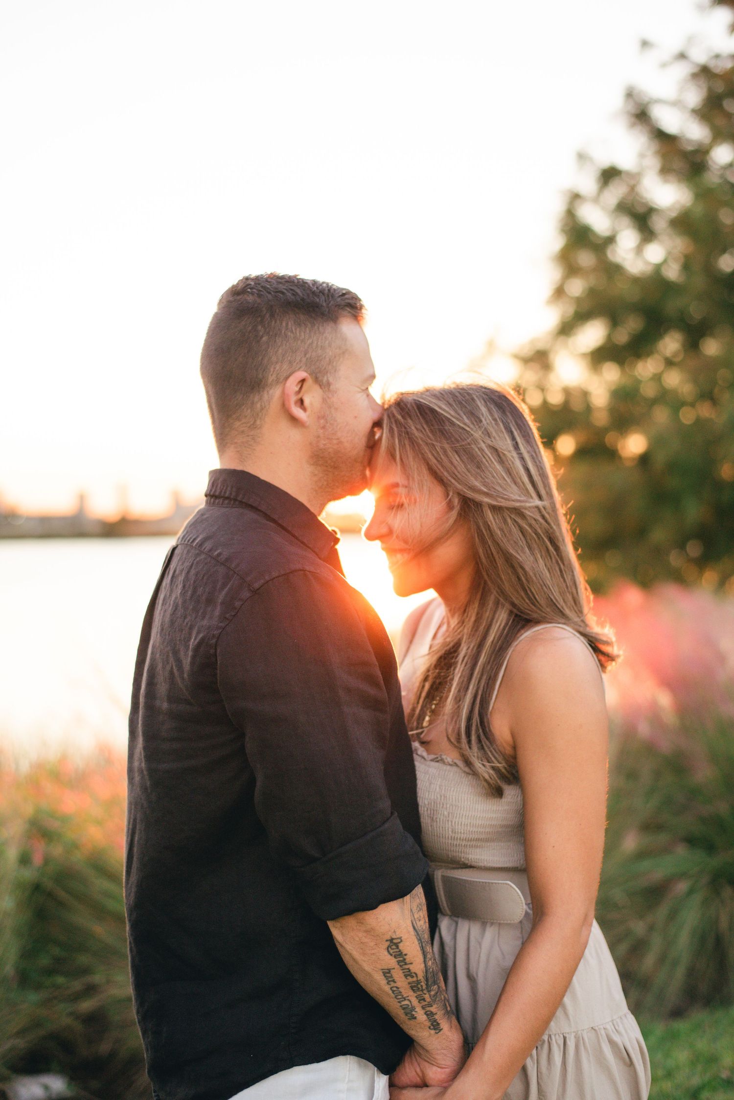 A couple shares an intimate moment at sunset by a lake with tall grass in the background.