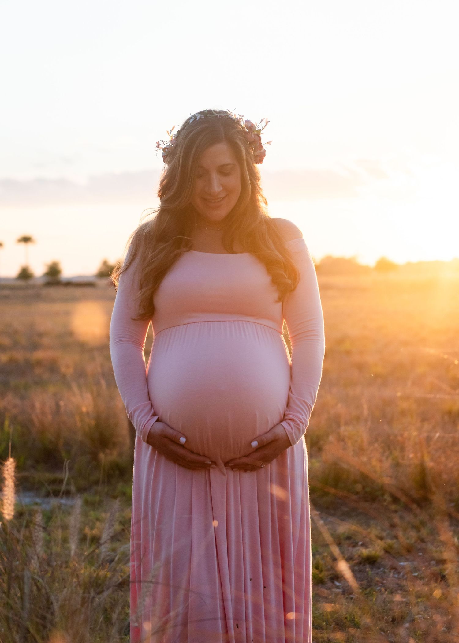 A pregnant person wearing a flower crown and pink dress poses in a field at golden hour.