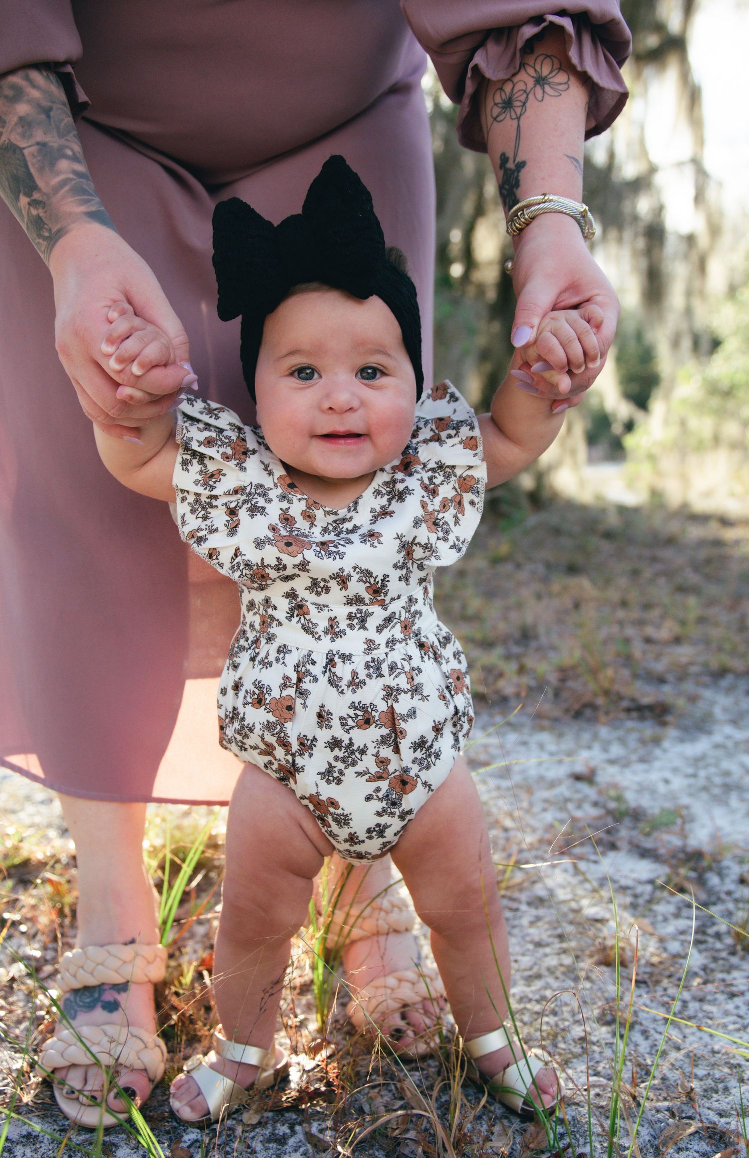 Infant wearing floral romper and black bow headband standing with support on sandy ground.
