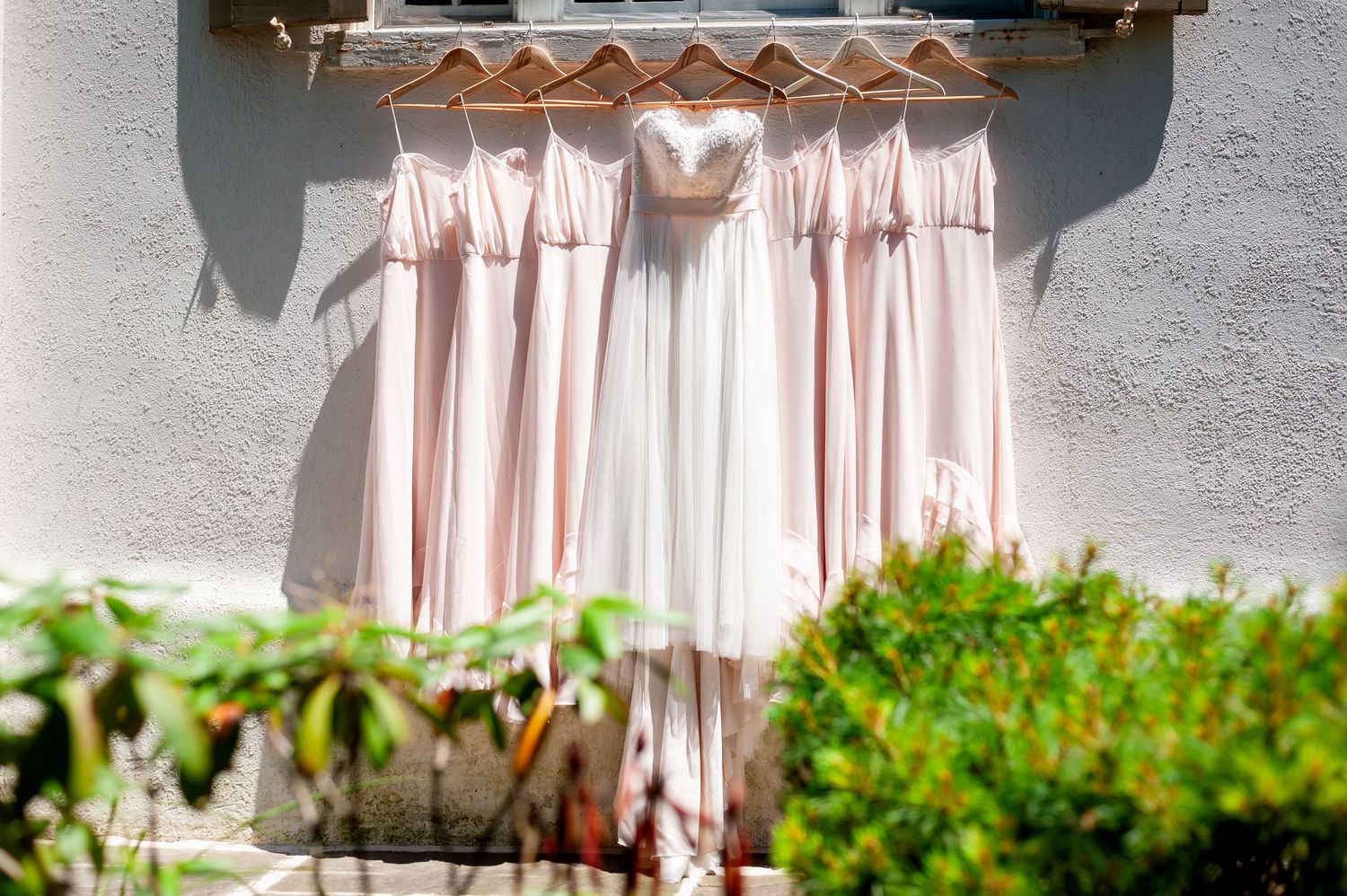 Blush pink bridesmaid dresses and white wedding gown hanging against grey wall.