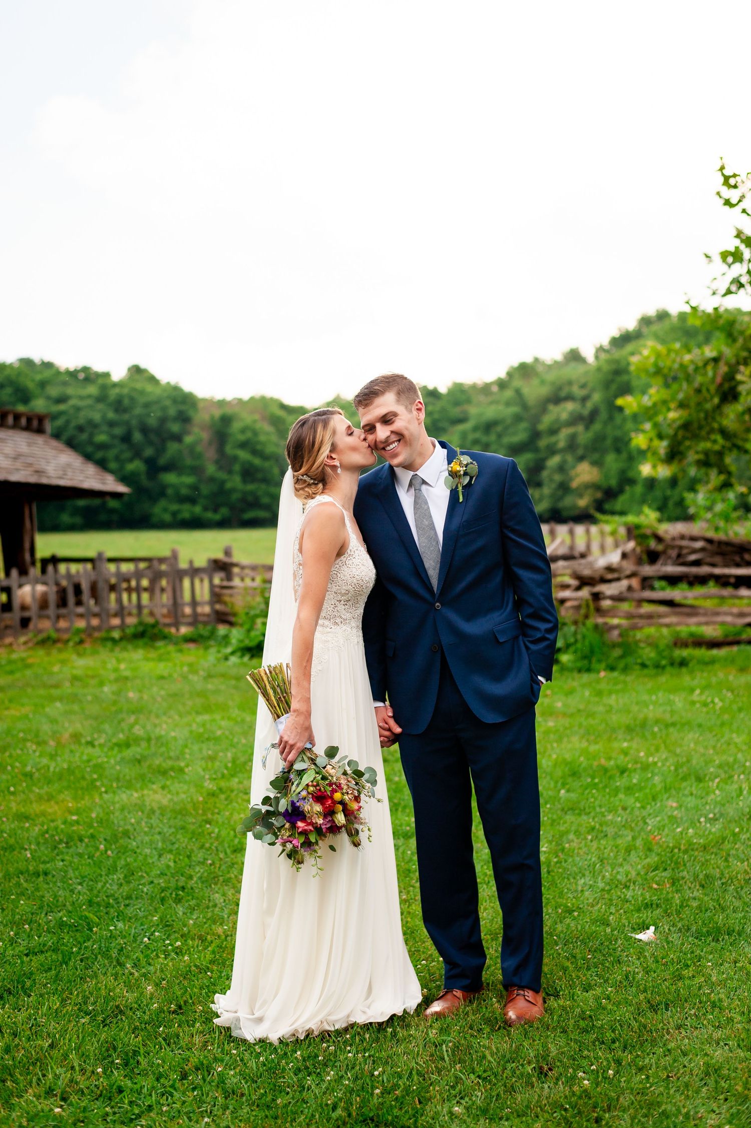 A couple in wedding attire stands on green lawn with wooden fence in background.