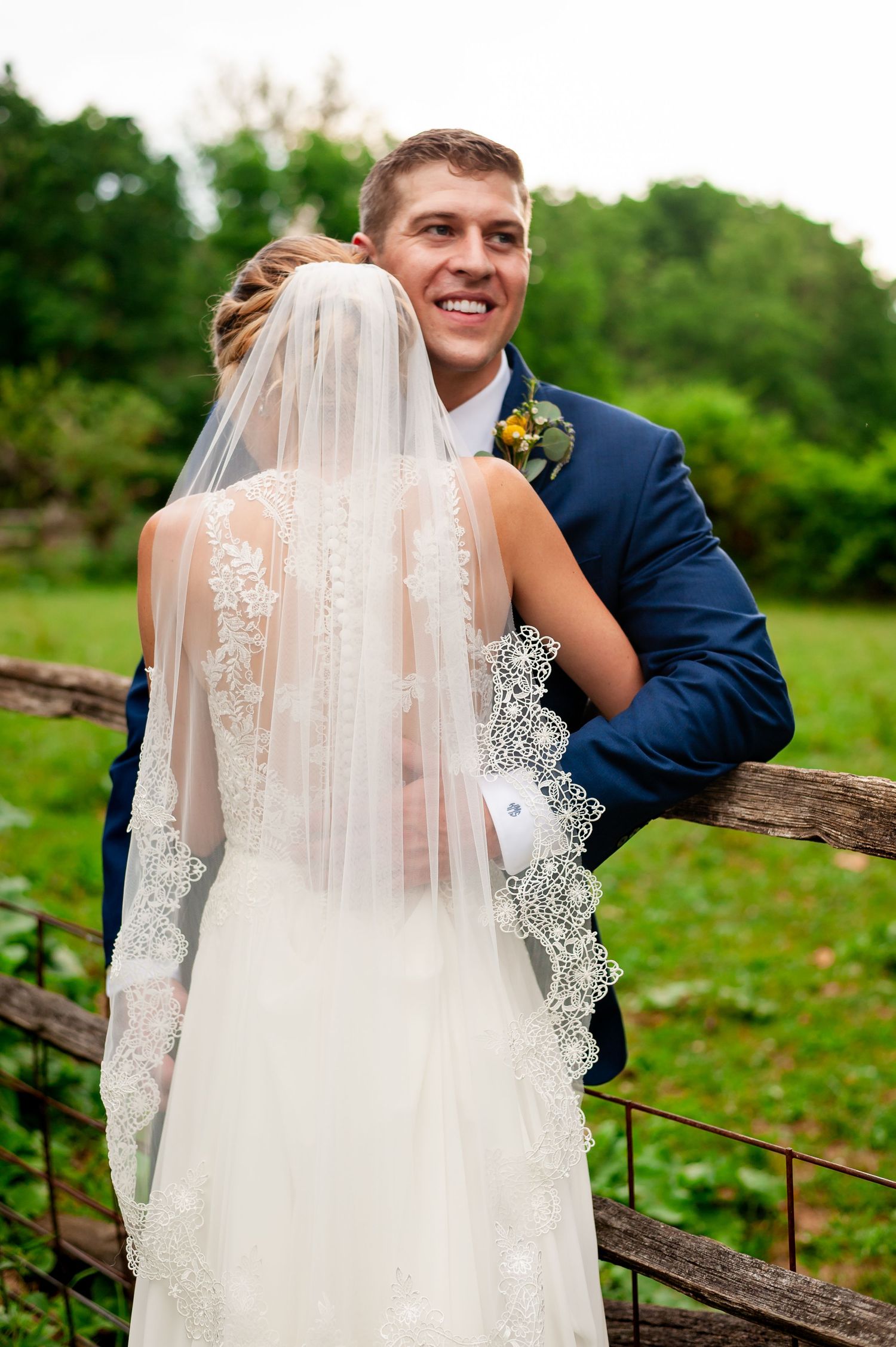Joyful wedding couple poses by a rustic wooden fence in an outdoor garden setting.