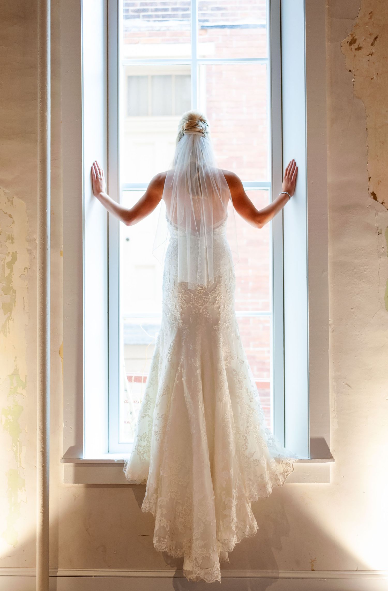 Silhouette of bride in lace gown and veil standing at sunlit window.